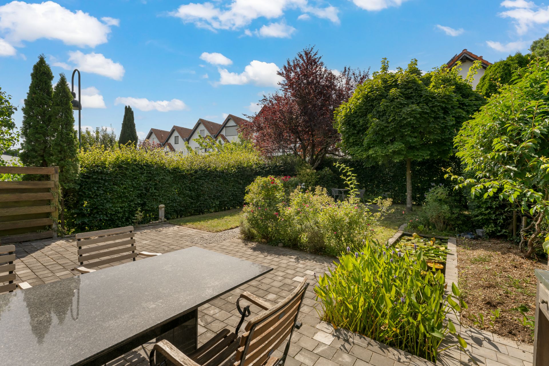 Gepflasterte Terrasse mit Tisch und Stühlen, umgeben von Garten und Bäumen unter blauem Himmel.