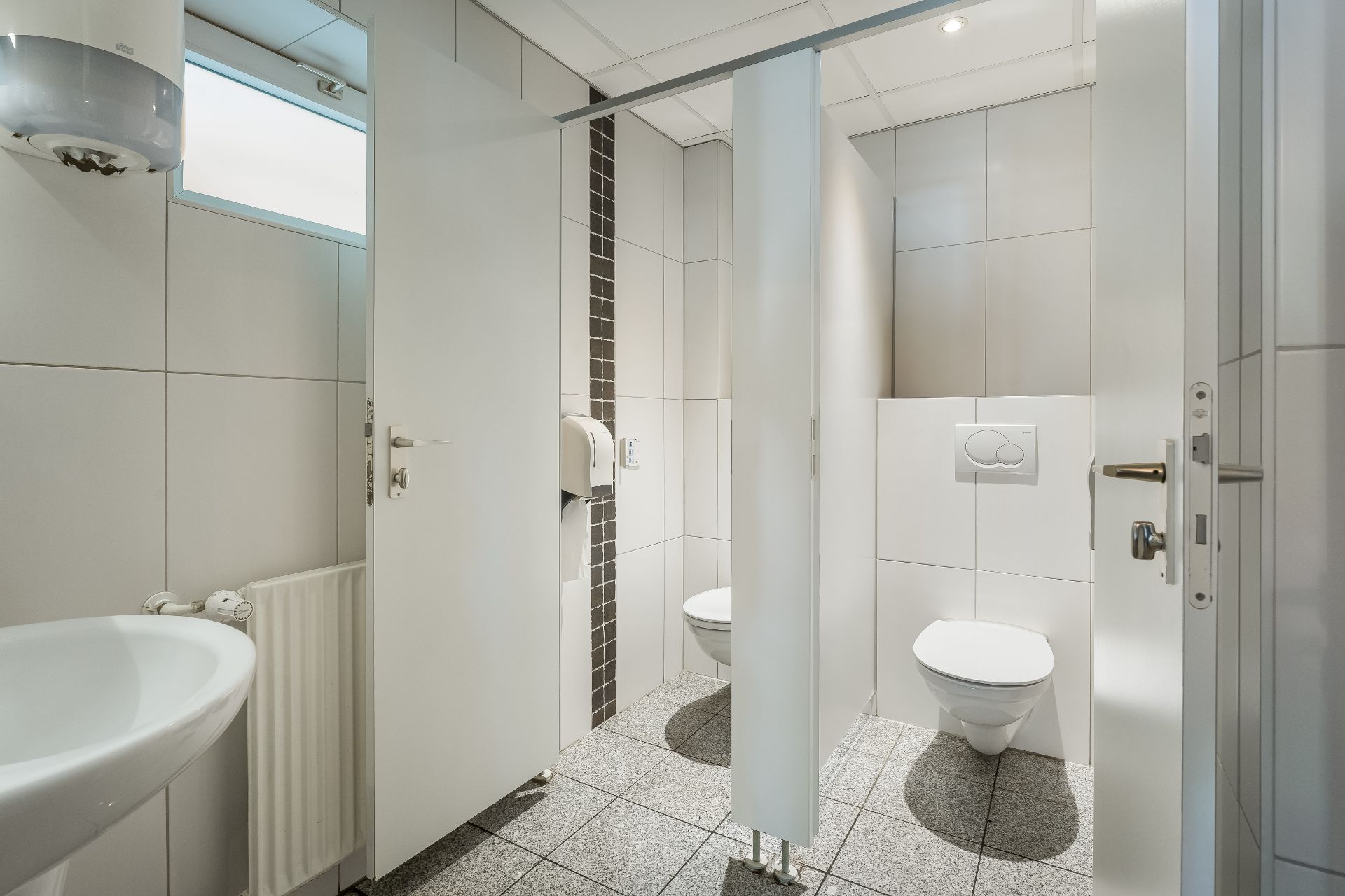 Heller Sanitärraum mit zwei Toilettenkabinen, Waschbecken und Papierhandtuchspender.