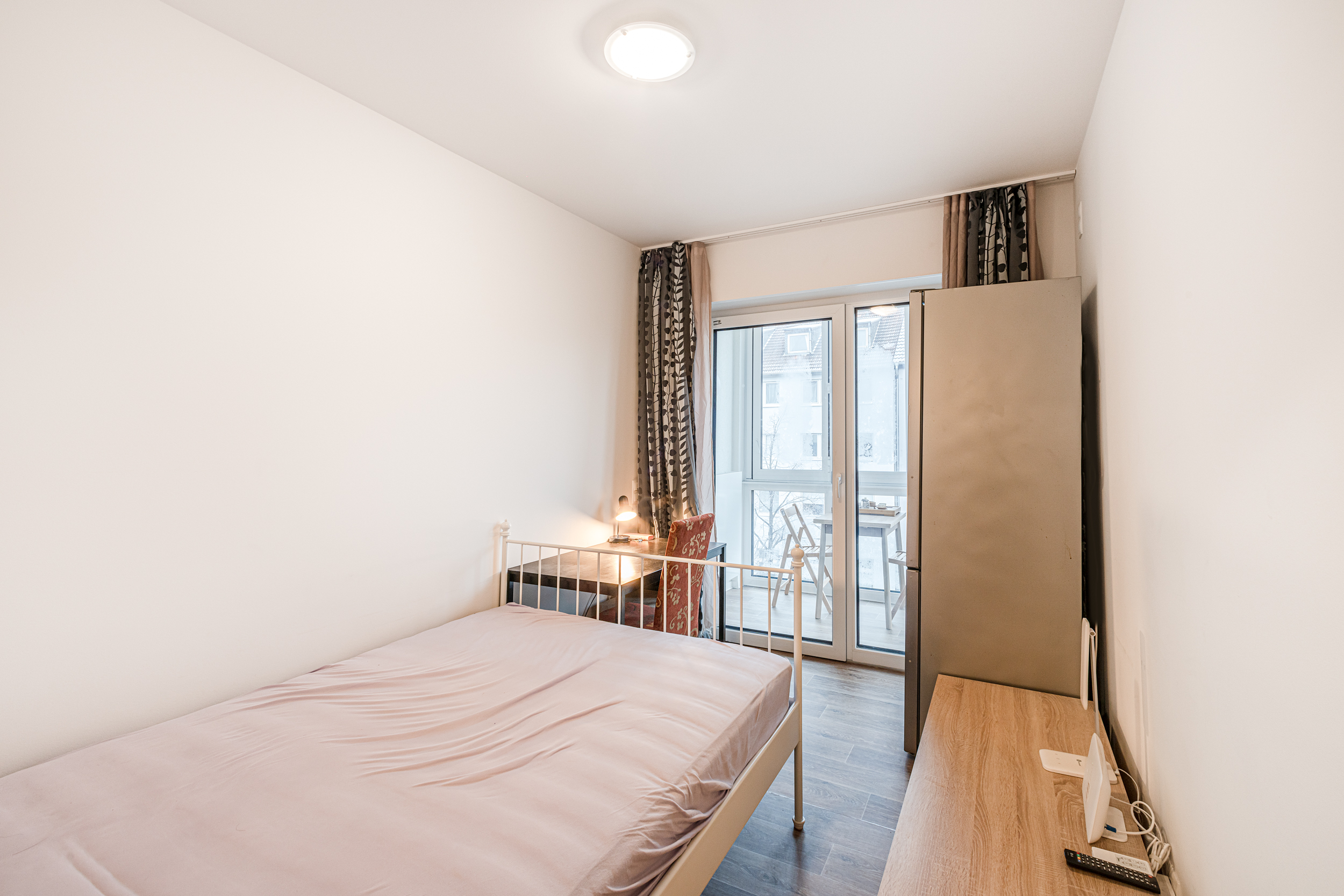 Helles Schlafzimmer mit Bett, Schreibtisch, Stuhl, Kleiderschrank und Zugang zu einem Balkon.