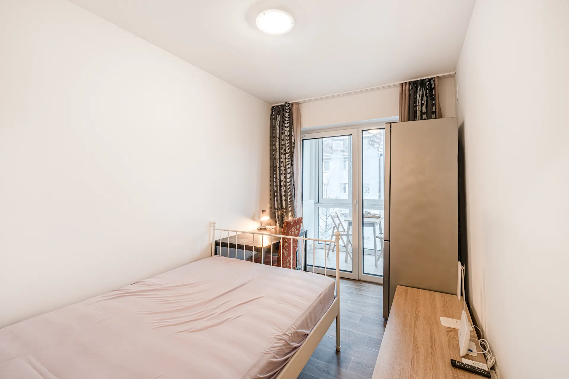 Helles Schlafzimmer mit Bett, Schreibtisch, Stuhl, Kleiderschrank und Zugang zu einem Balkon.