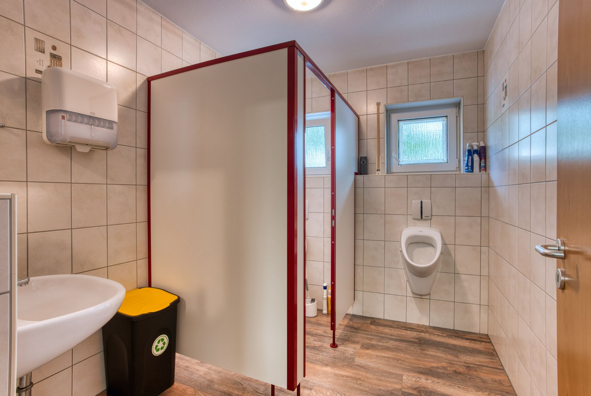 Gefliester Waschraum mit Urinal, Waschbecken, Abfallbehälter und Kabine, zwei kleine Fenster an der Wand.