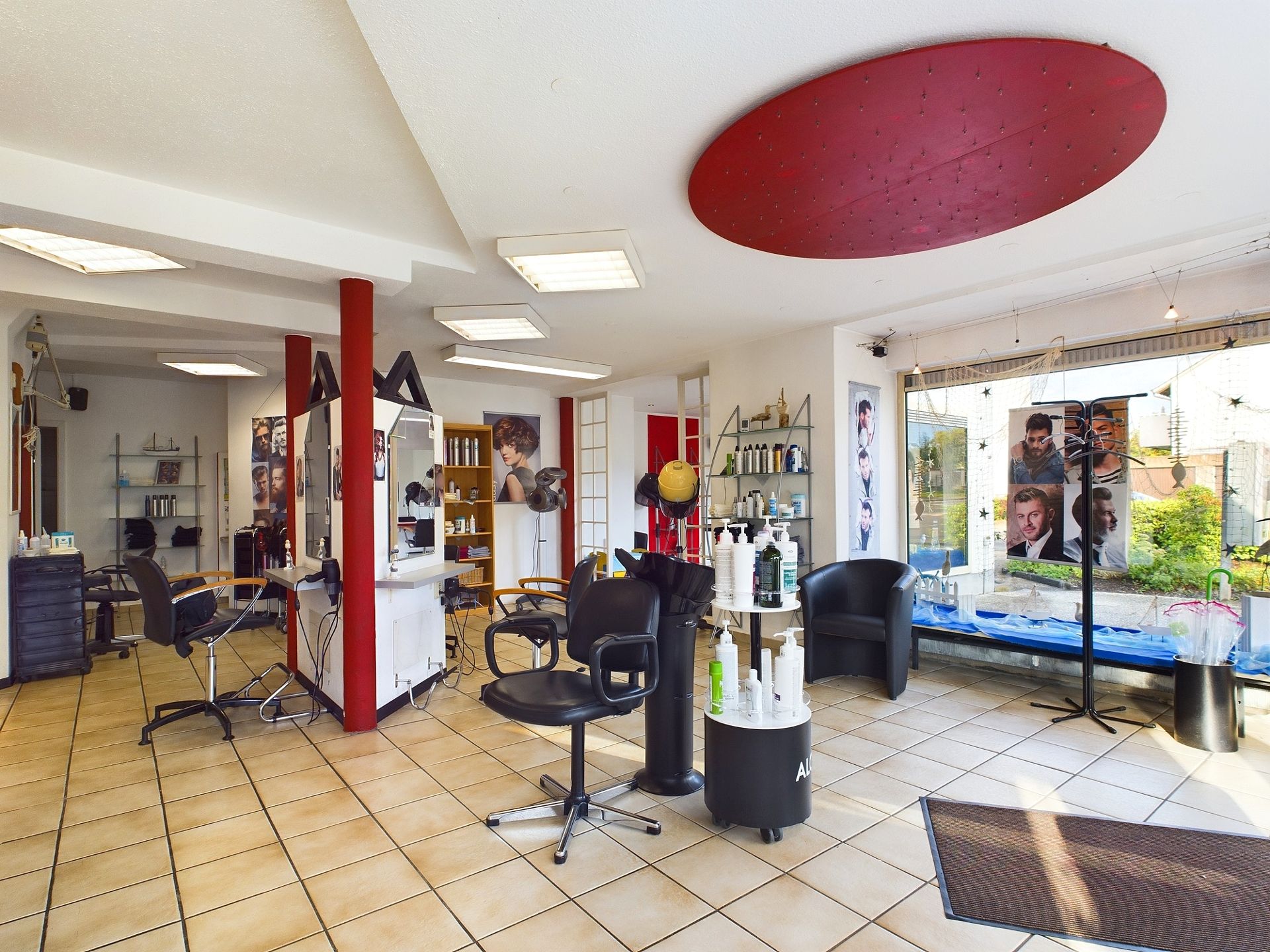 Ein heller Friseursalon mit Friseurstühlen, Spiegeln und Produkten, Fliesenboden und großen Fenstern.