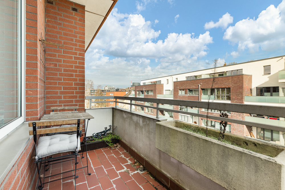 Balkon mit Tisch und Stuhl, Blick auf moderne Wohngebäude und blauen Himmel mit Wolken.