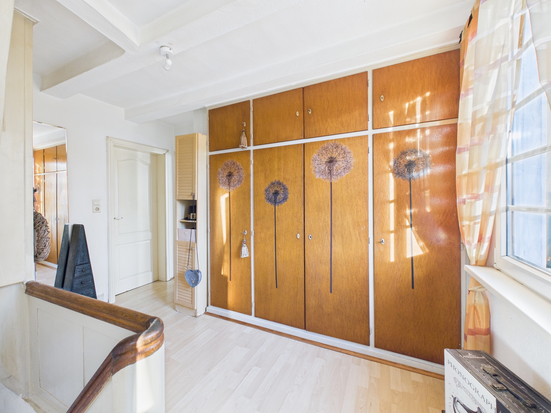 Flur mit großen Holzschränken, auf denen Pusteblumen aufgemalt sind, und einem Fenster mit Vorhang.