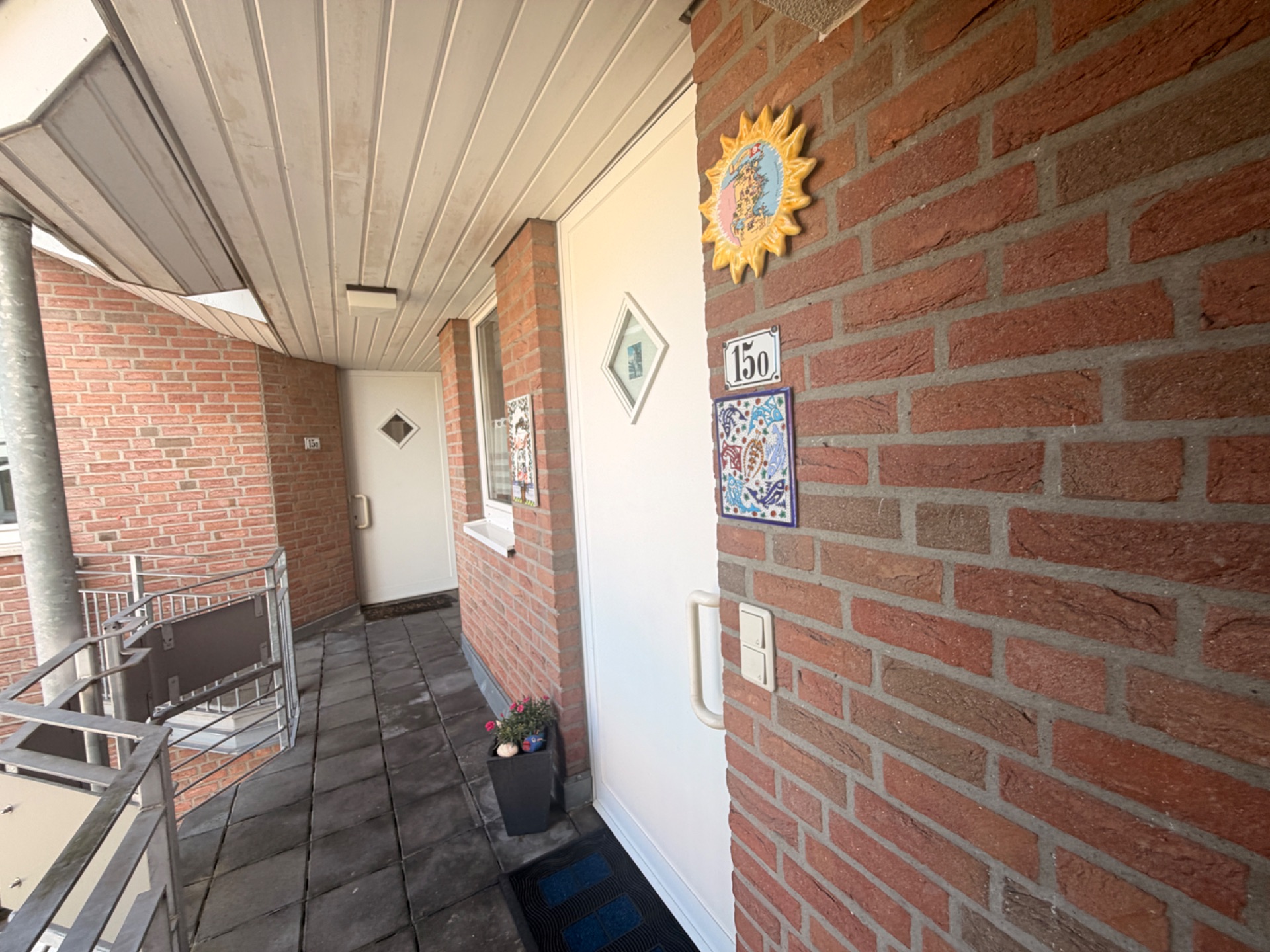 Eingangsbereich mit zwei weißen Türen, roter Ziegelwand und Blumenkasten auf einem Balkon.