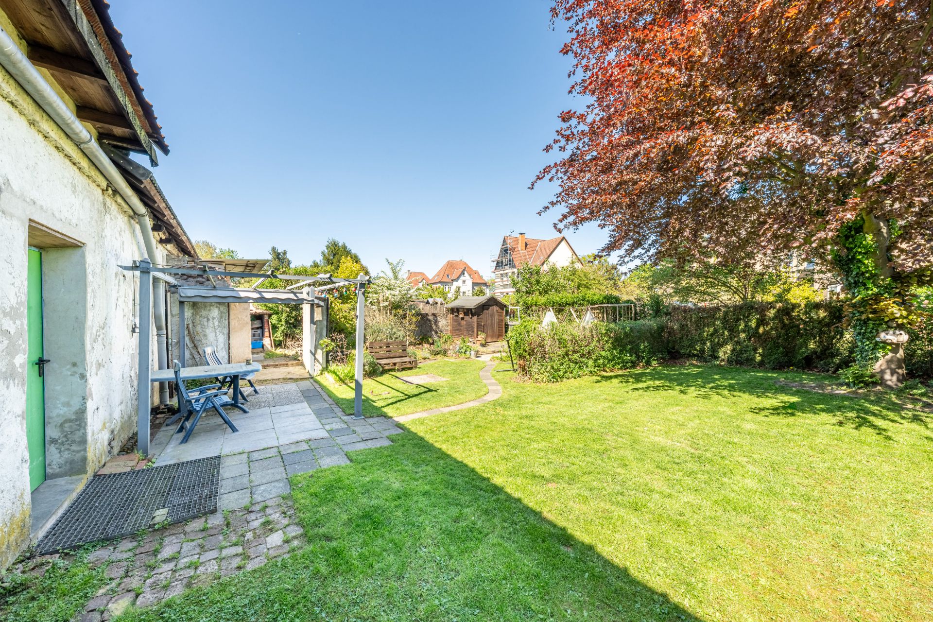 Grüner Garten mit Rasen, Baum, Terrasse und Gartenmöbeln neben einem weißen Gebäude.