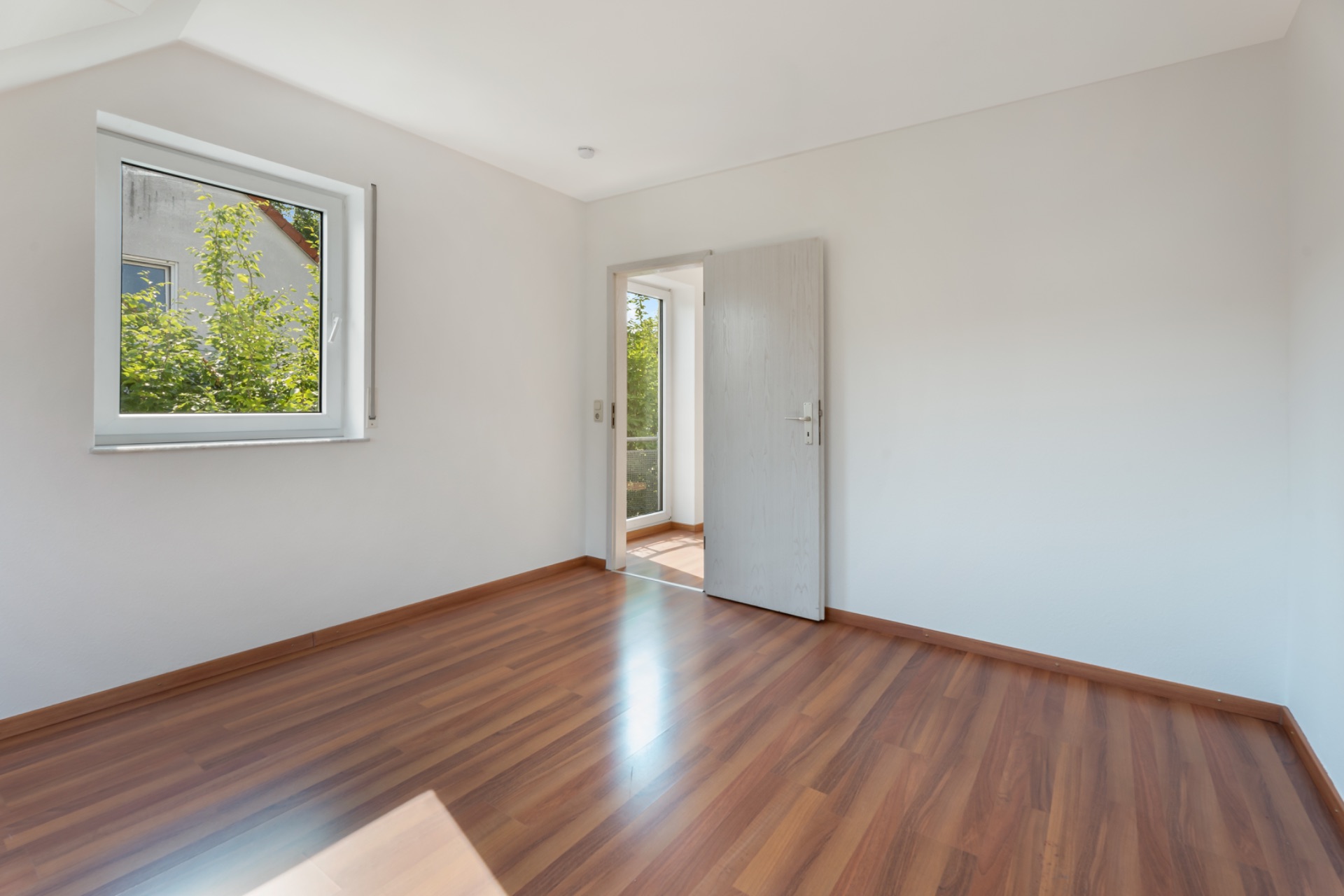 Helles, leeres Zimmer mit Holzfußboden, Fenster und offener Tür zum Balkon.