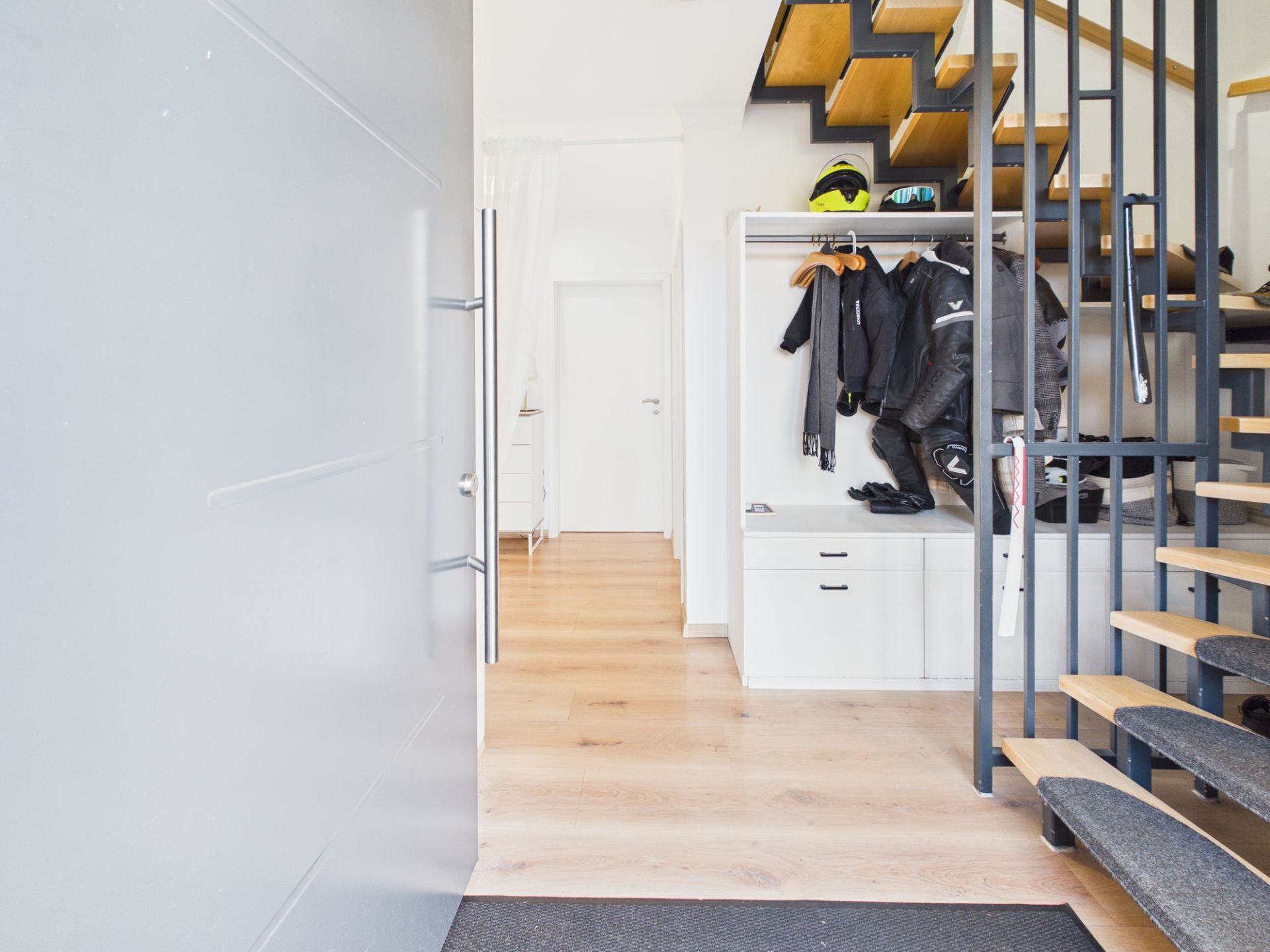 Heller Flur mit Holztreppe, Garderobe und Jacken, Blick auf eine weiße Tür.