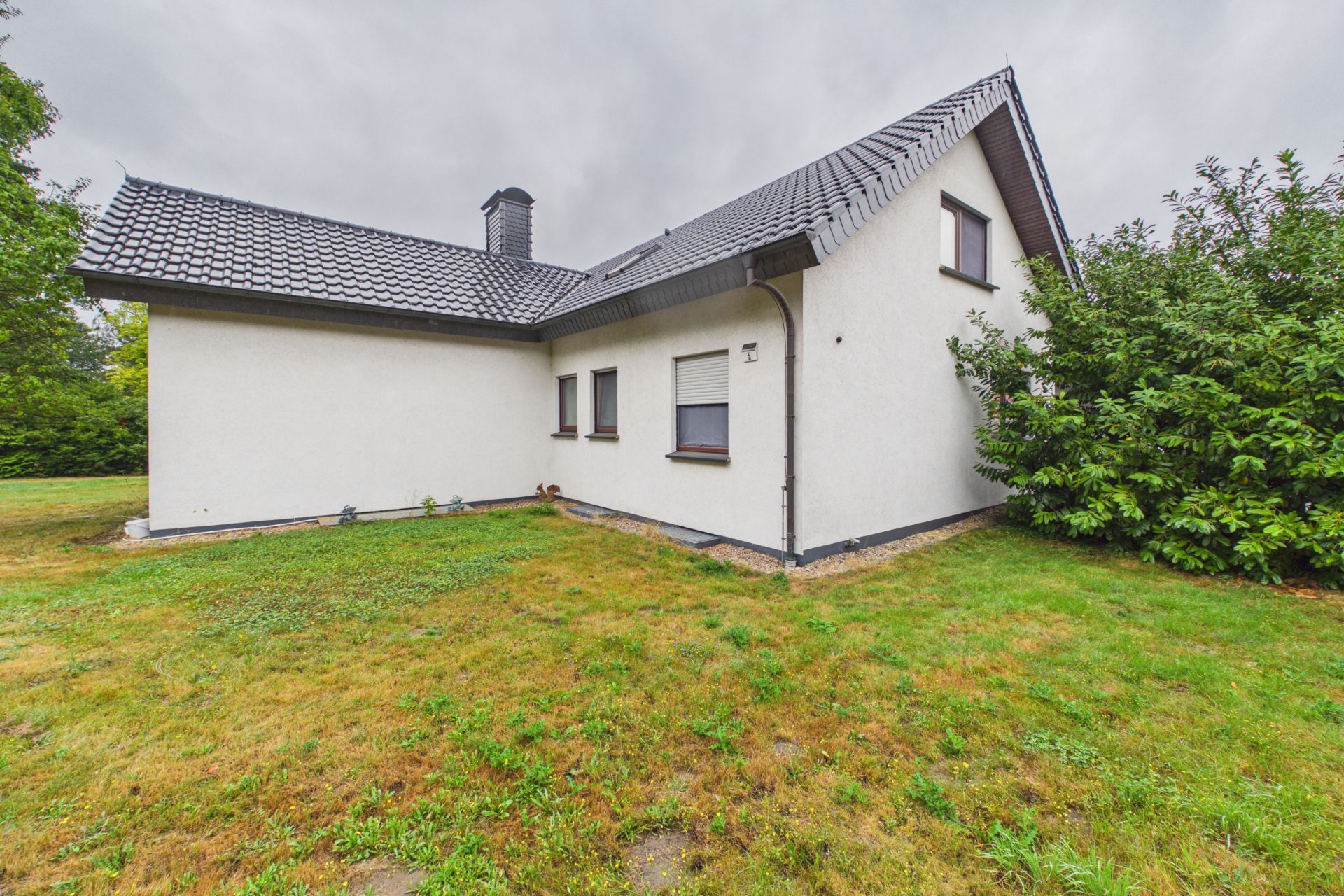 Einfamilienhaus mit weißer Fassade, dunklem Dach und Garten, rechts ein großer Strauch.