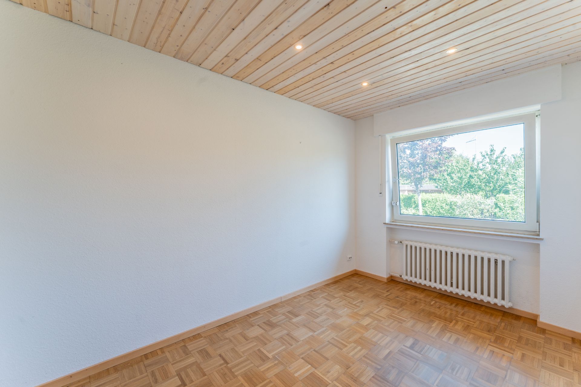 Helles, leeres Zimmer mit Holzdecke, Parkettboden und großem Fenster mit Blick ins Grüne.