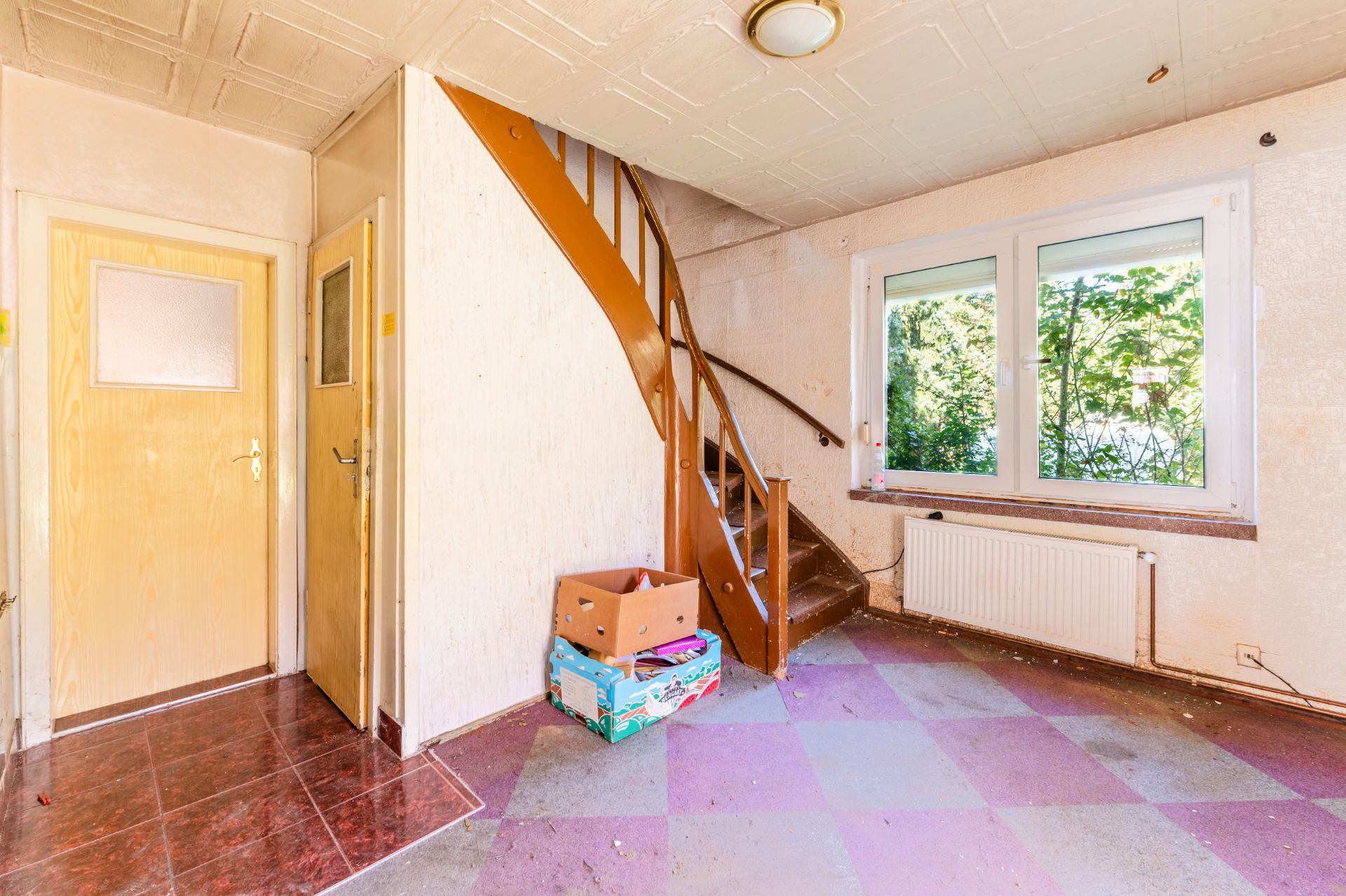 Eingangsbereich mit Holztreppe, zwei Türen, großem Fenster und Kartons auf dem Boden.