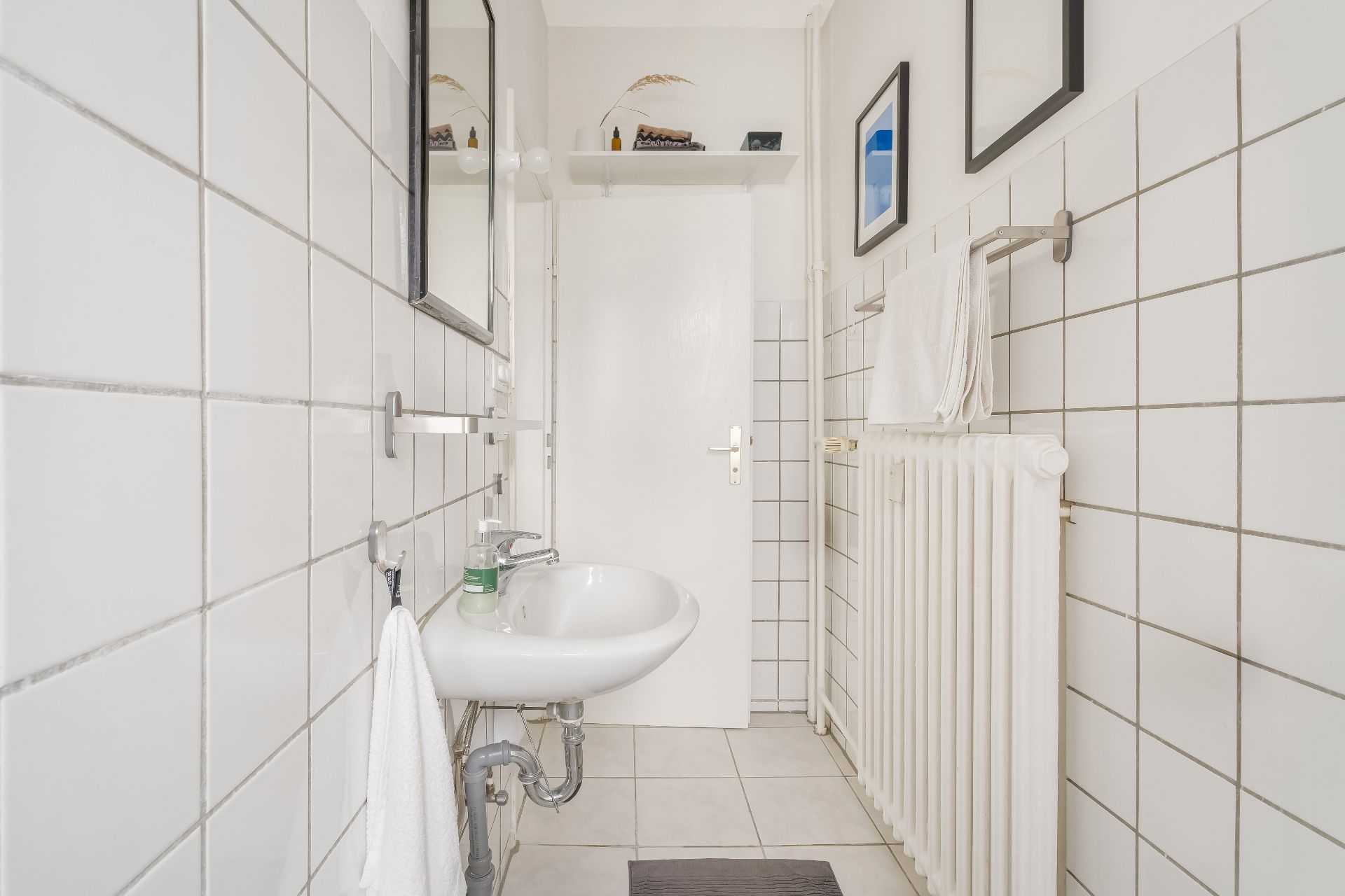 Helles Badezimmer mit weißen Fliesen, Waschbecken, Handtüchern und Heizkörper.