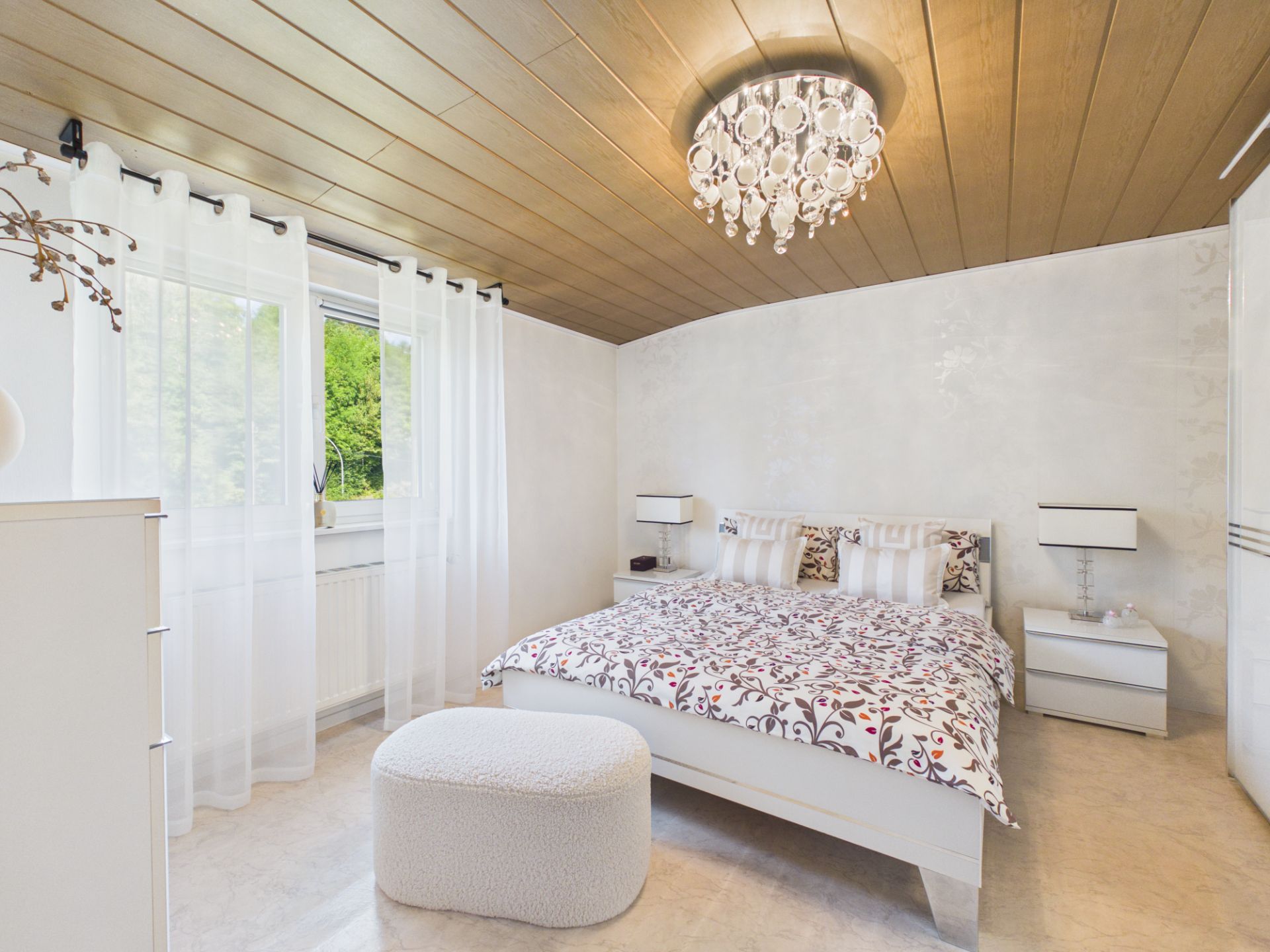 Helles Schlafzimmer mit Doppelbett, Nachttischen, weißem Hocker und Kronleuchter.