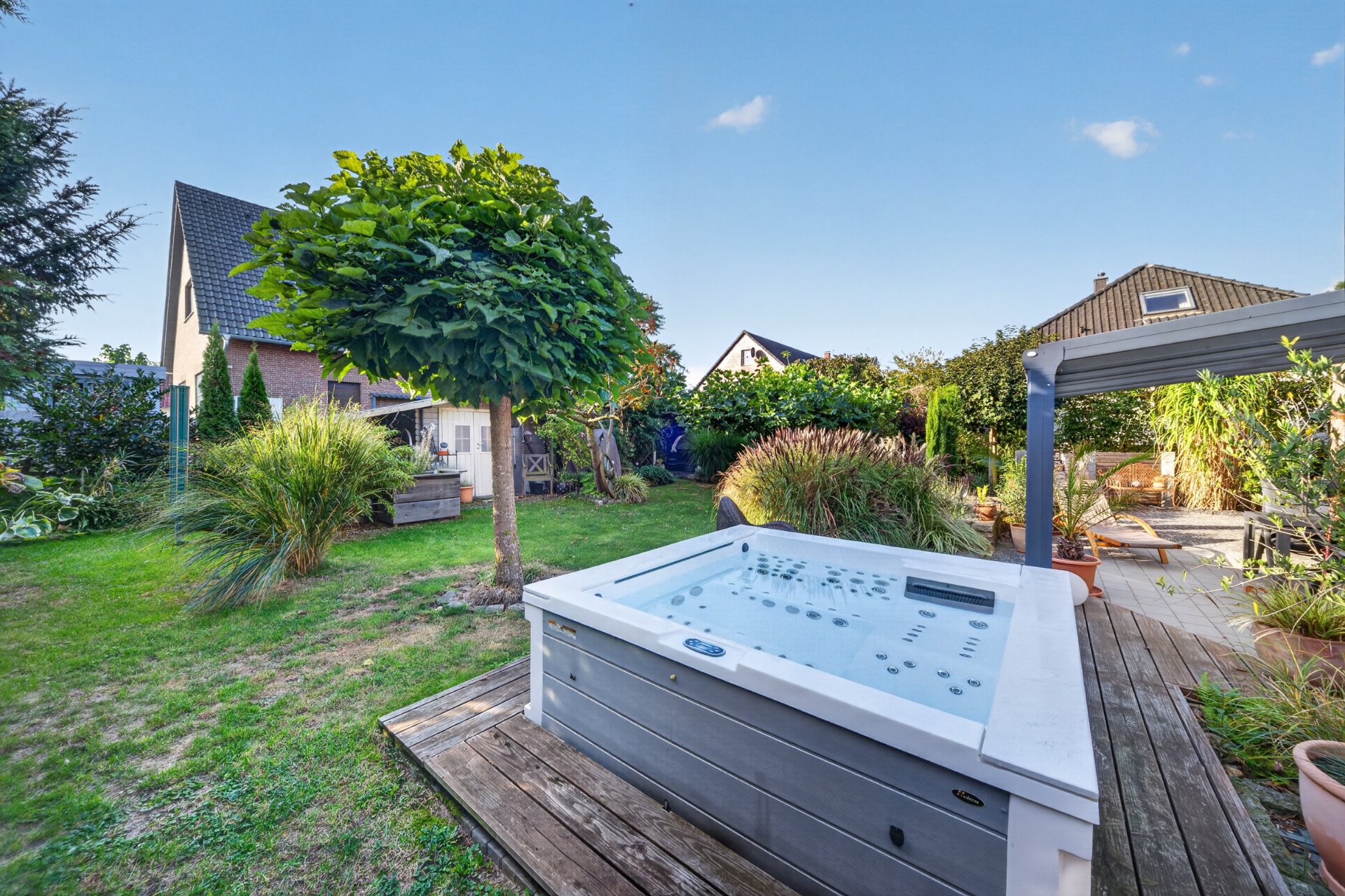 Garten mit Whirlpool, Holzterrasse, Baum und verschiedenen Pflanzen, Haus im Hintergrund.