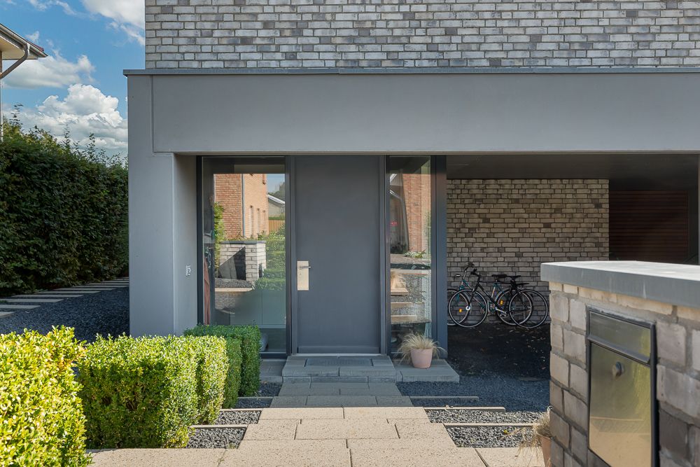 Moderne Hauseingangstür mit Glas, davor Pflanzentopf und Hecken, rechts zwei Fahrräder unter Carport.