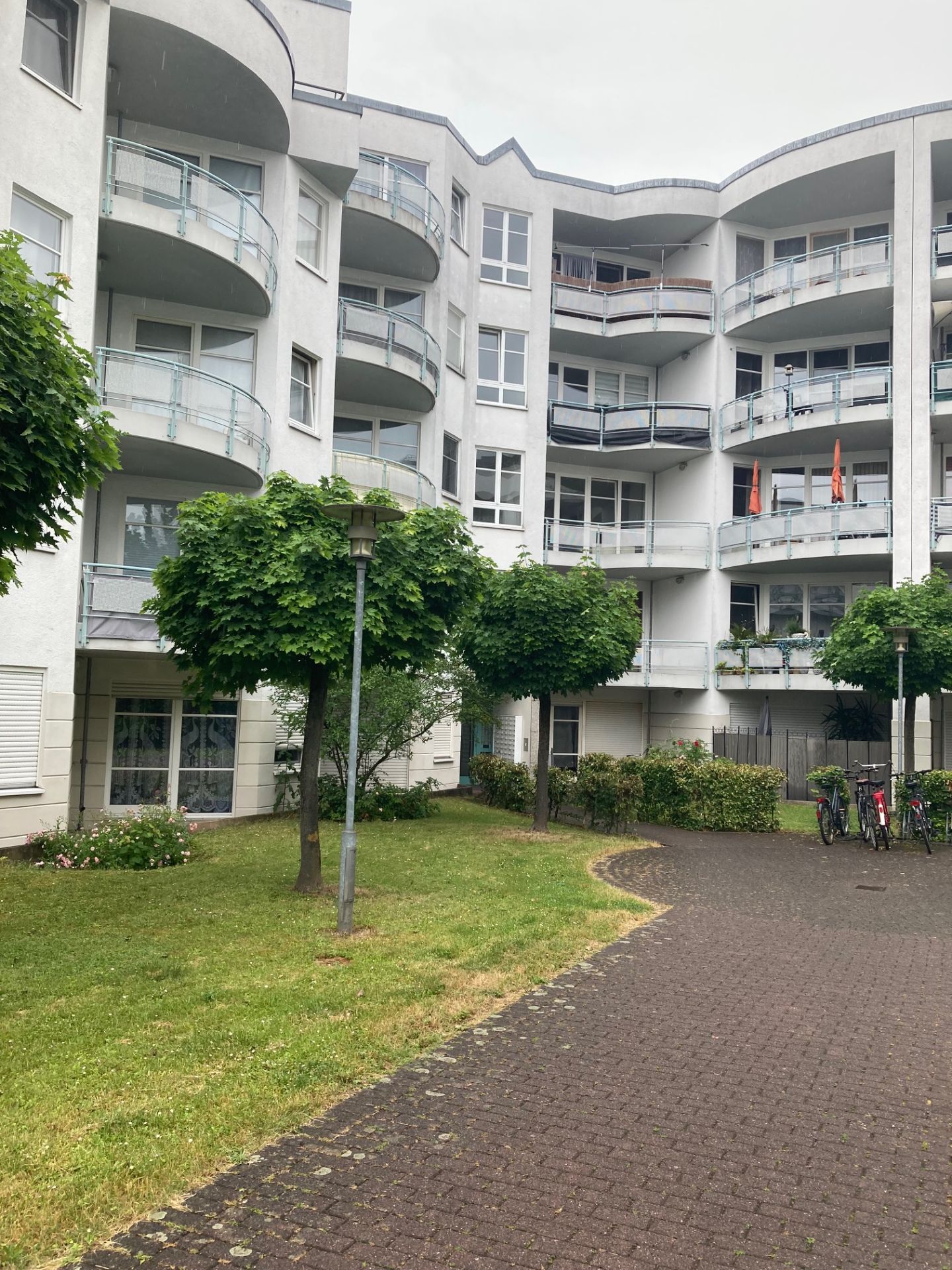 Mehrstöckiges Wohnhaus mit Balkonen, Bäumen, Rasen und gepflastertem Weg im Vordergrund.