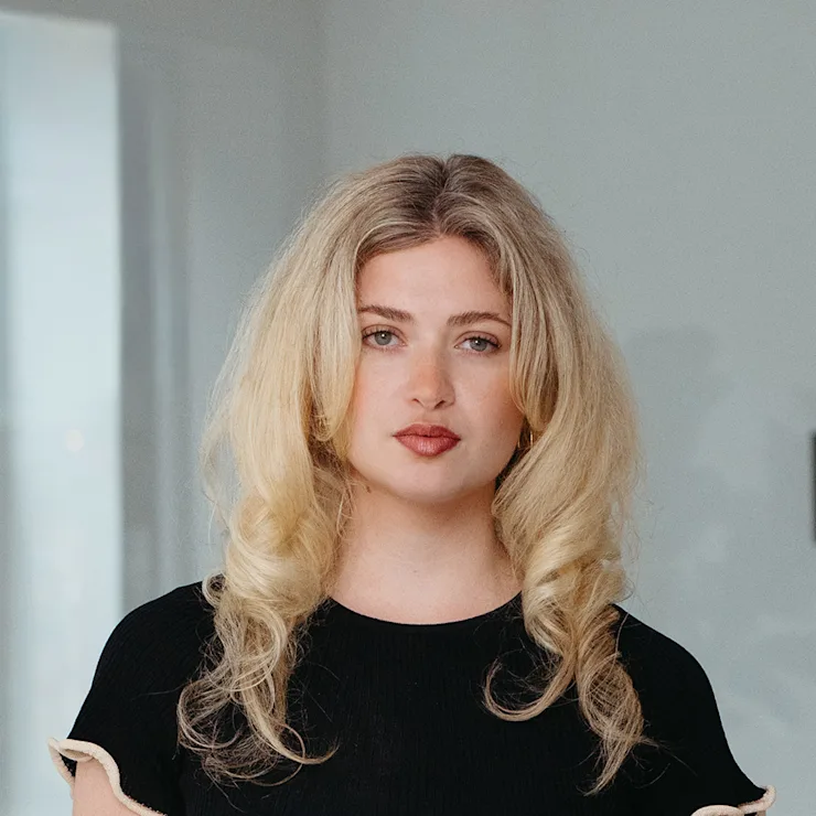 Eine lächelnde Frau mit langen, blonden Haaren trägt einen beigen Blazer und ein schwarzes Shirt.