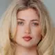 Eine lächelnde Frau mit langen, blonden Haaren trägt einen beigen Blazer und ein schwarzes Shirt.