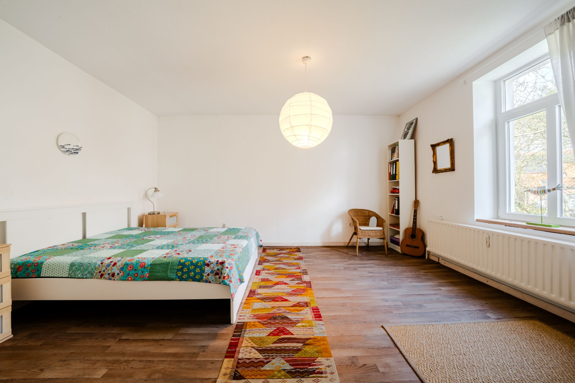 Großes Schlafzimmer mit Bett, bunter Decke, Teppich, Fenster, Regal und Gitarre.