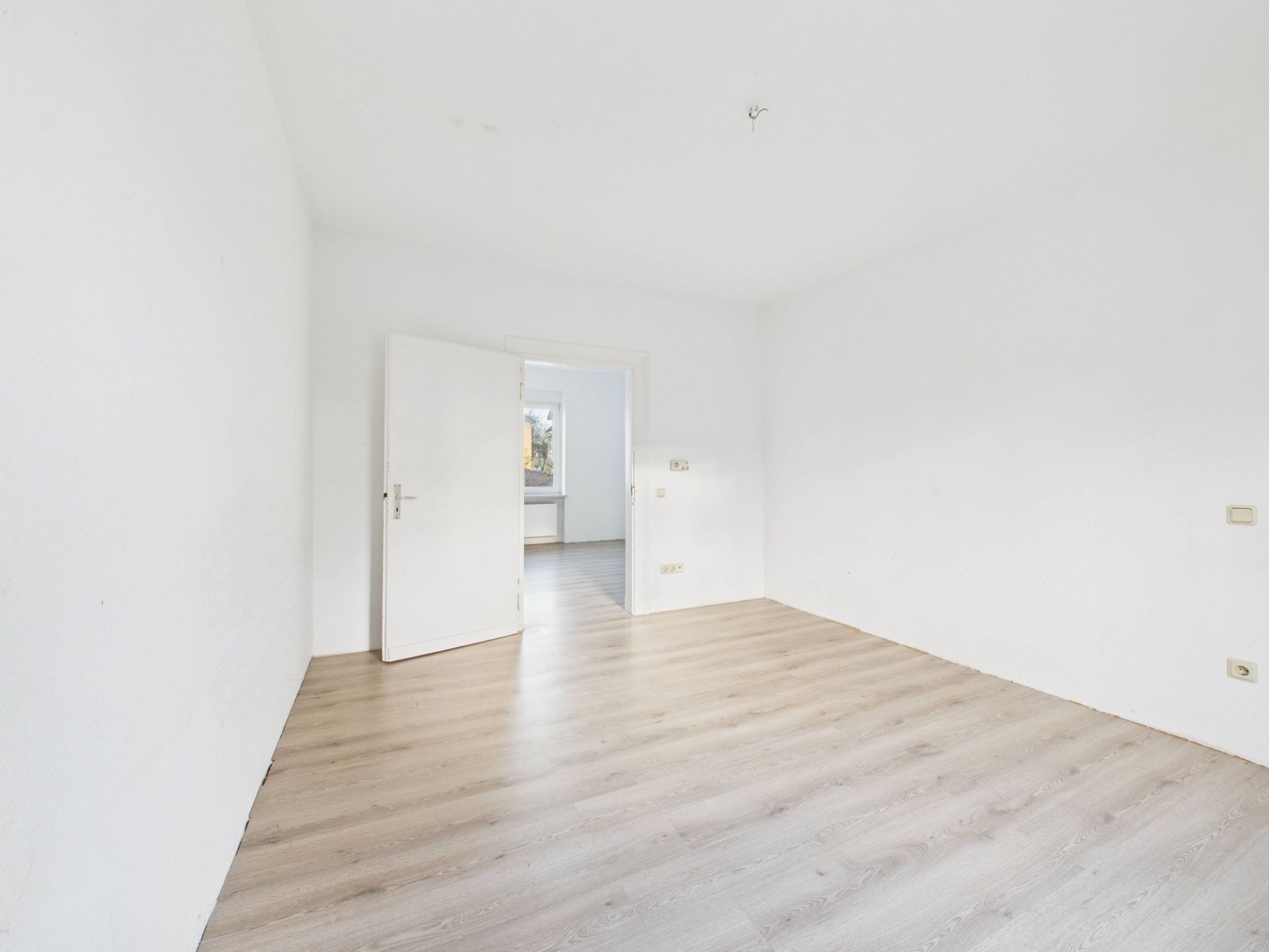 Heller leerer Raum mit weiß gestrichenen Wänden und hellem Holzboden, offene Tür zum nächsten Zimmer.