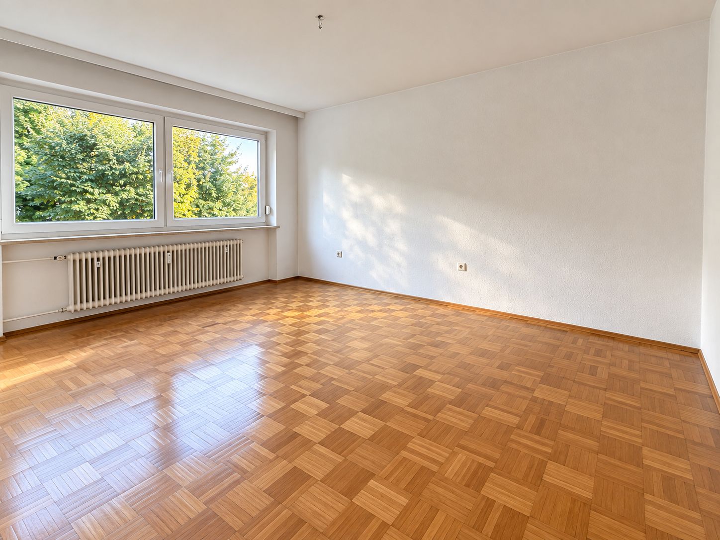 Leerer Raum mit Parkettboden, weißer Wand und großem Fenster mit Blick auf Bäume.