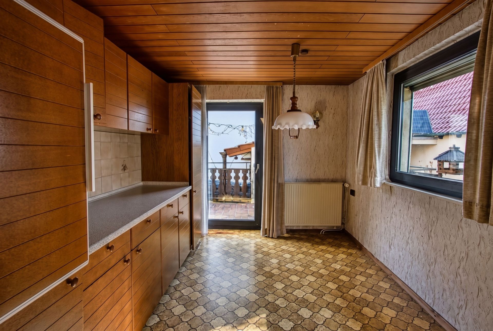 Küche mit Holzschränken, Fenster, Tür zum Balkon und gemustertem Boden.