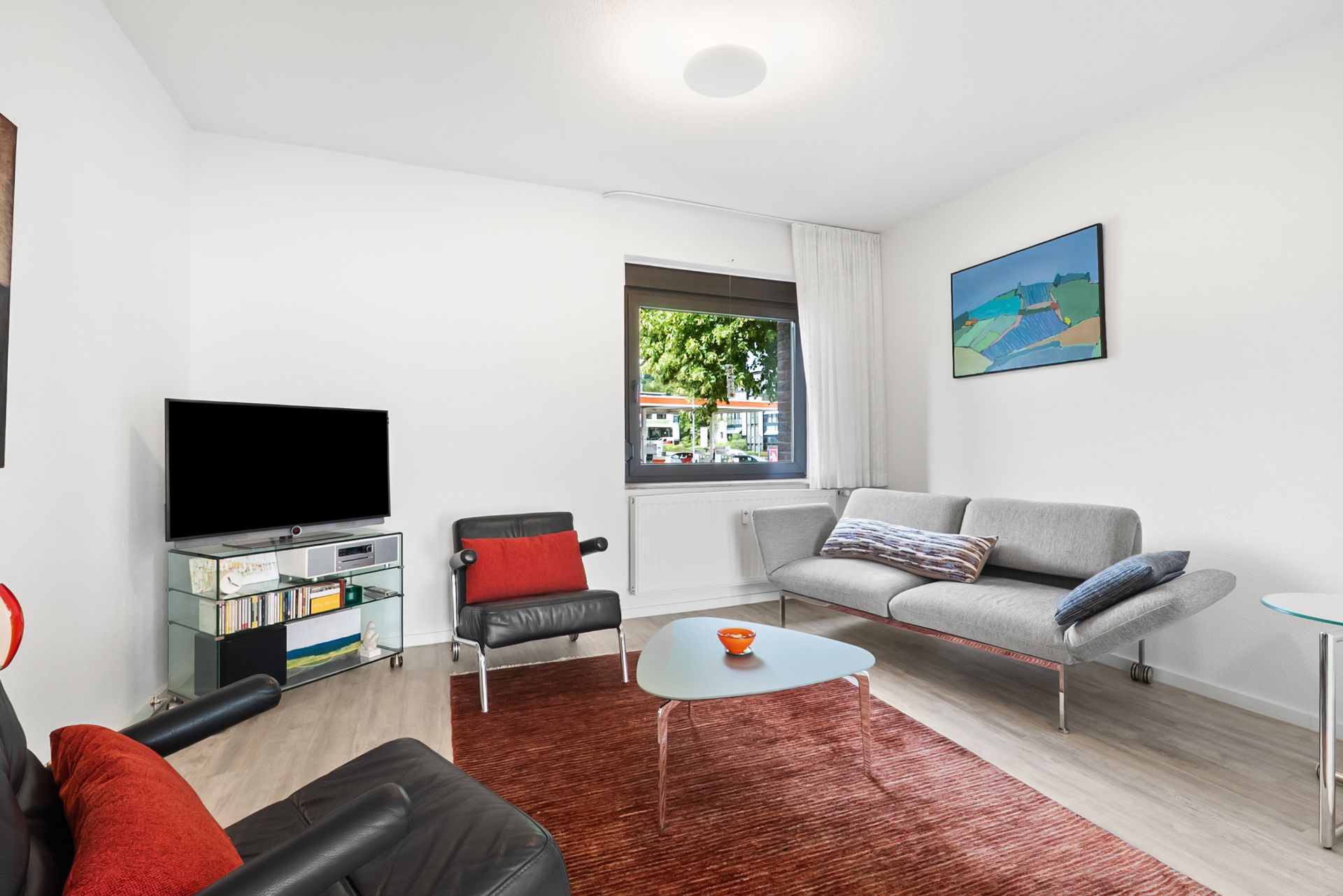 Wohnzimmer mit grauem Sofa, Sesseln, TV, rotem Teppich und Fenster mit Blick nach draußen.