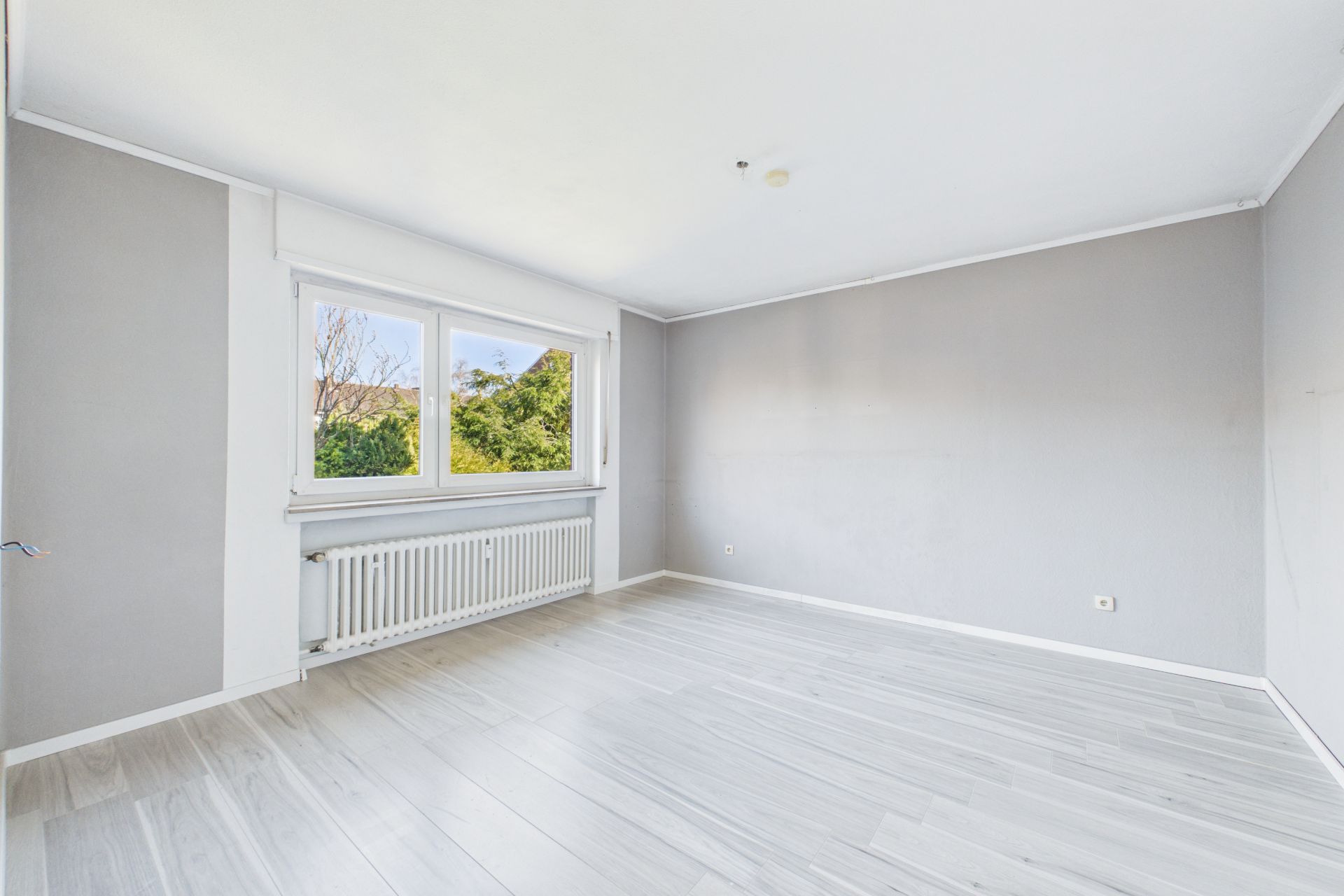 Helles, leeres Zimmer mit großem Fenster, grauen Wänden und hellem Boden.