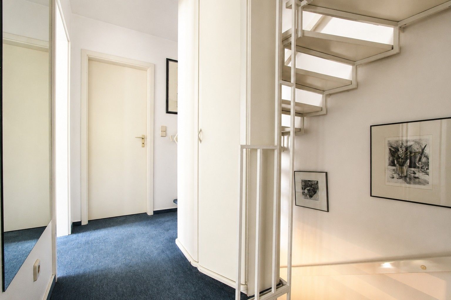 Heller Flur mit blauer Auslegware, weißer Treppe und weißen Wänden mit Bildern.