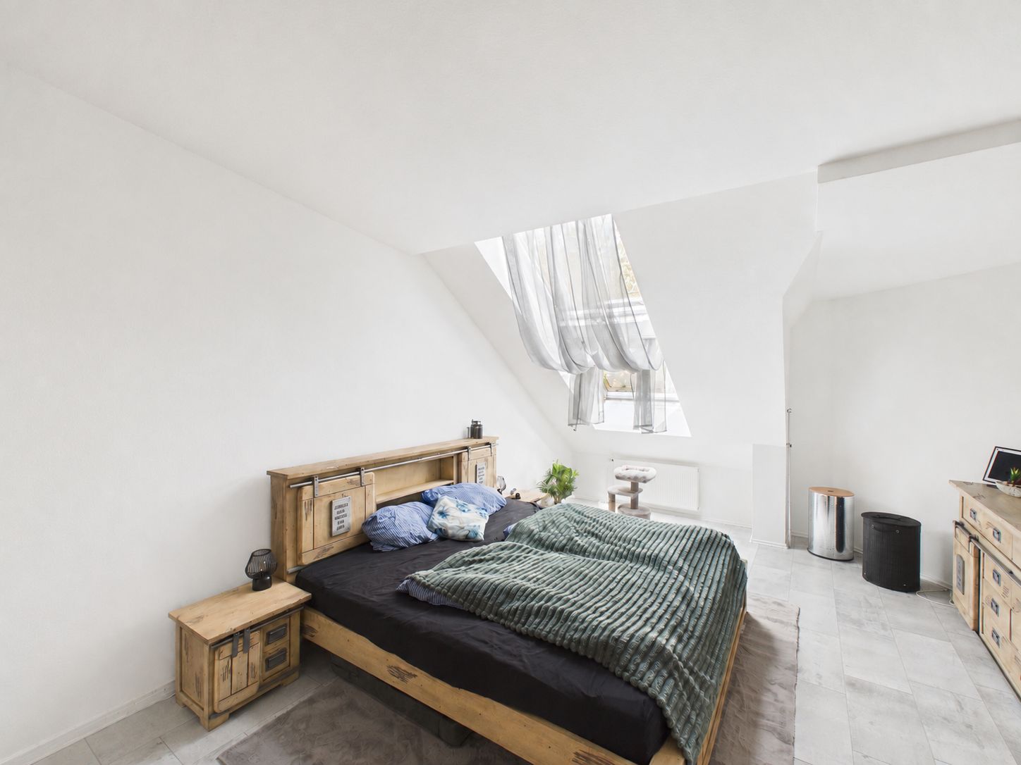 Helles Schlafzimmer mit Holzbett, Nachttisch und schrägem Fenster mit Vorhang.