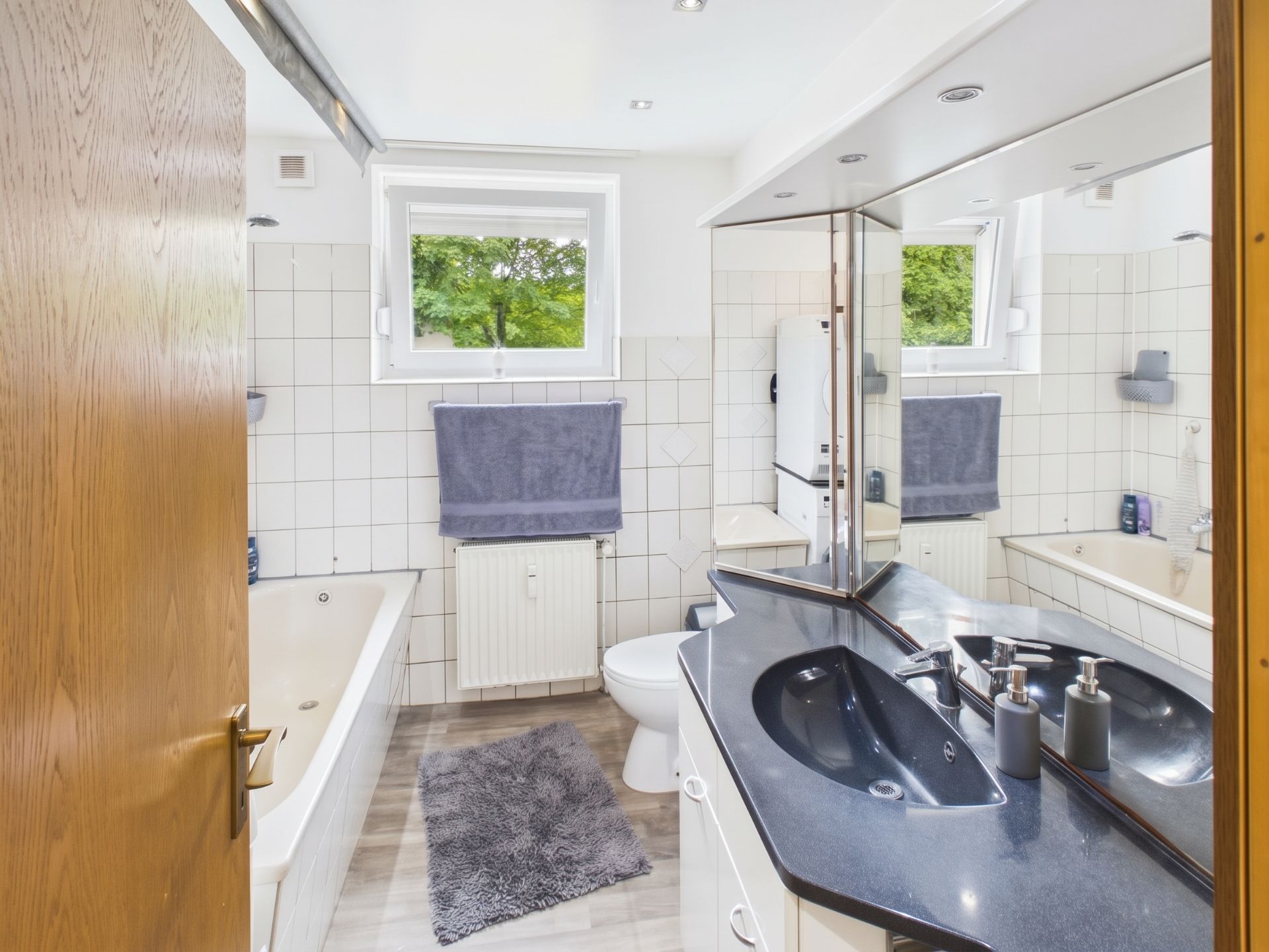 Helles Badezimmer mit Badewanne, Spiegel, schwarzem Waschbecken und Fenster.