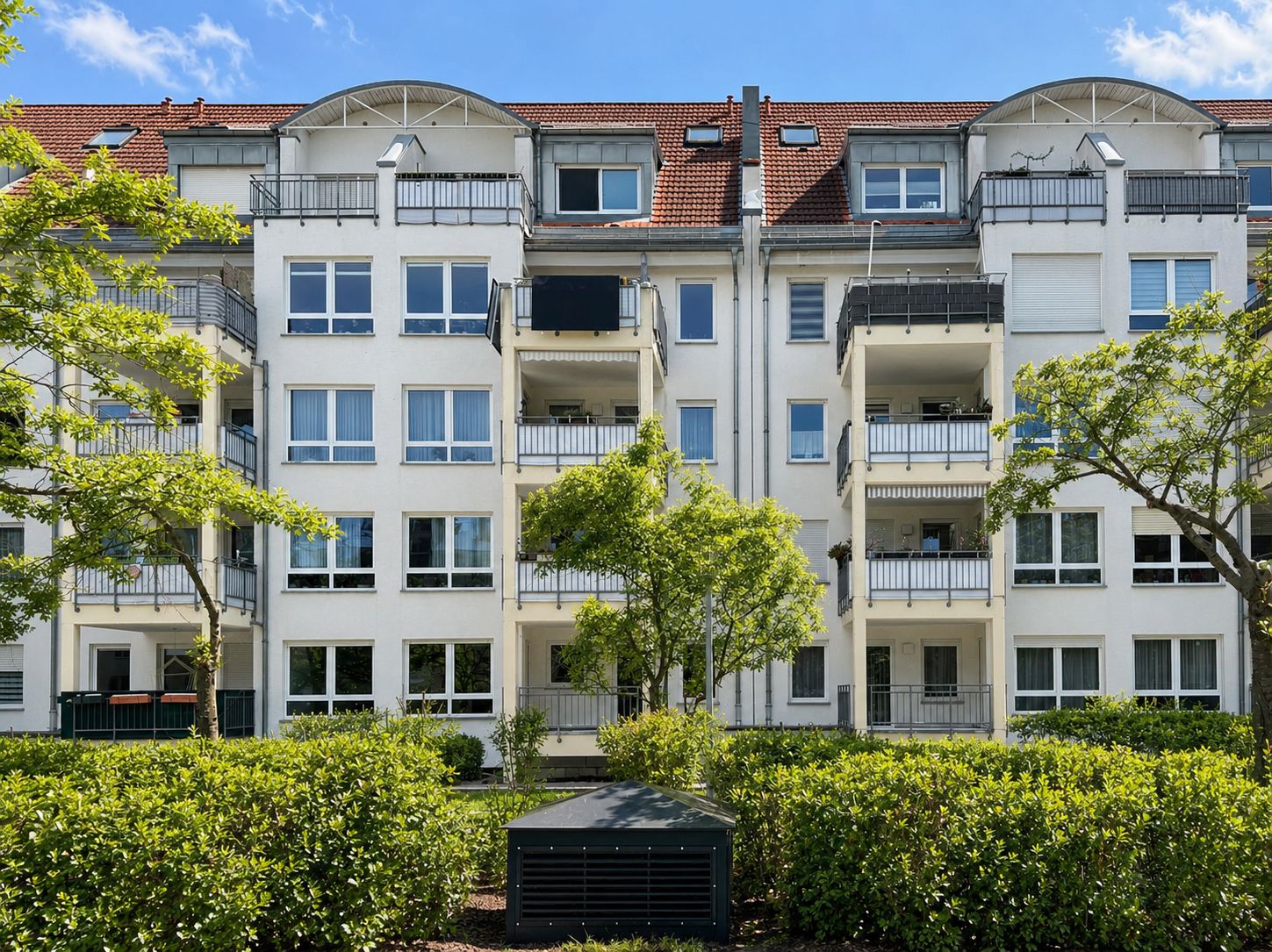 Mehrstöckiges Wohnhaus mit Balkonen, Bäumen und Sträuchern im Vordergrund.