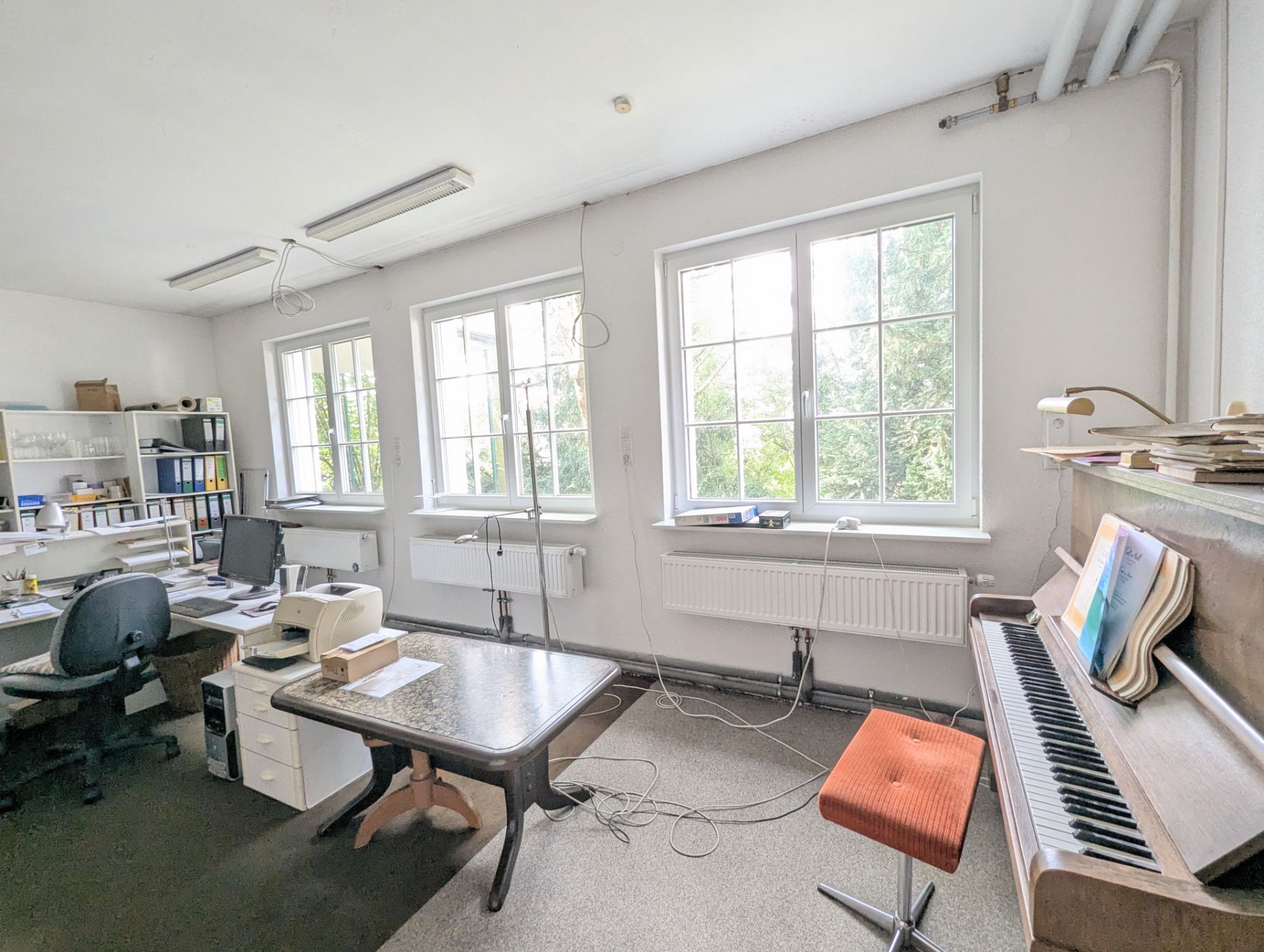 Helles Arbeitszimmer mit Schreibtischen, Regalen, Klavier und großen Fenstern.