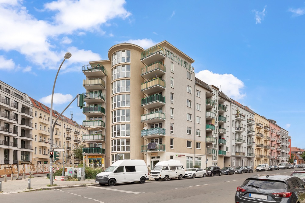Mehrstöckiges Wohnhaus mit Balkonen an einer Straßenecke, geparkte Autos davor.