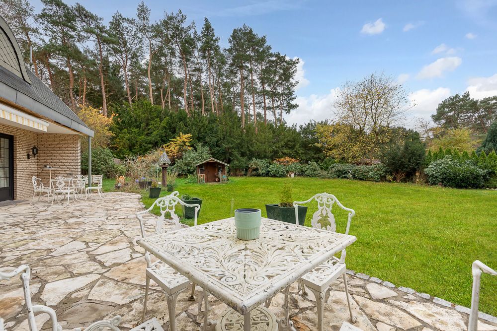 Garten mit Terrasse, weißen Metallstühlen und Tisch, Blick auf Rasen und Bäume im Hintergrund.