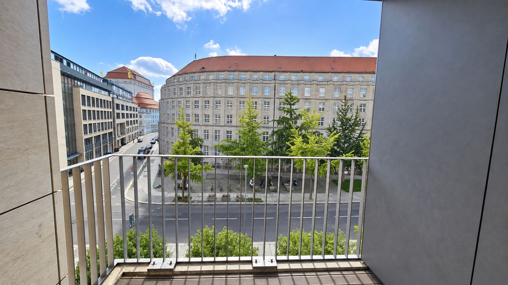 Blick von einem Balkon auf eine Straße mit Bäumen und gegenüberliegendem großes Gebäude.