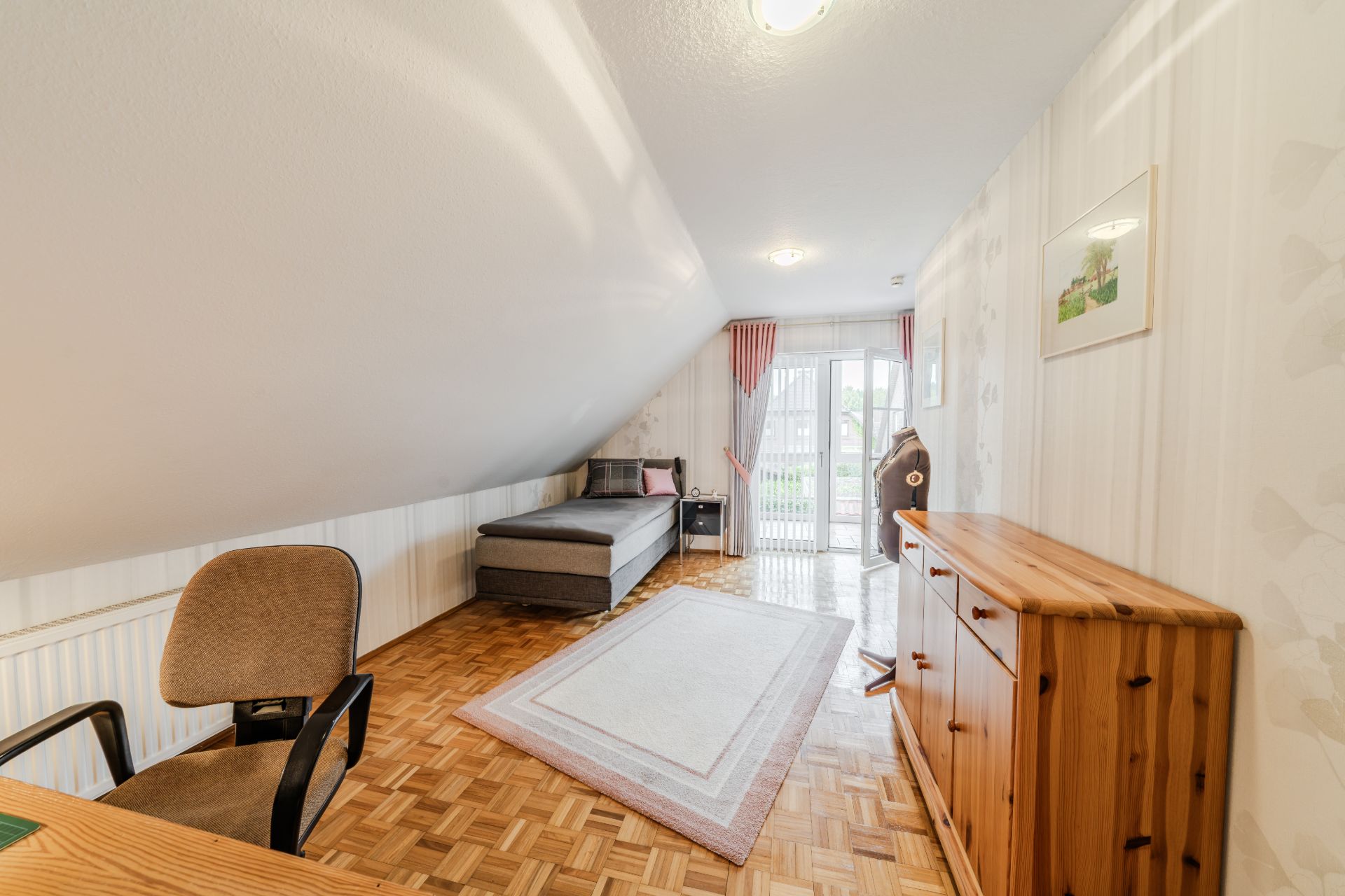 Zimmer mit Schreibtisch, Stuhl, Schlafsofa, Teppich und Kommode unter einer Dachschräge.