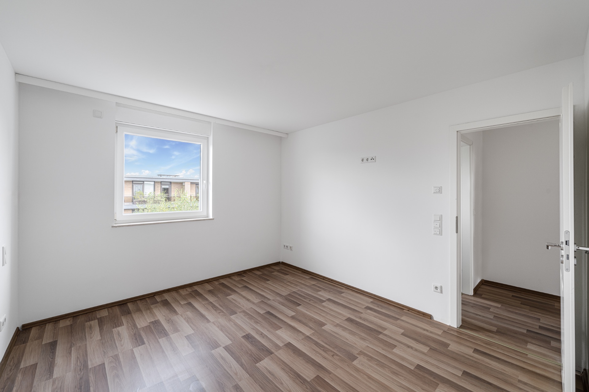 Leerer Raum mit braunem Laminatboden, weißem Fenster und weißer Tür.