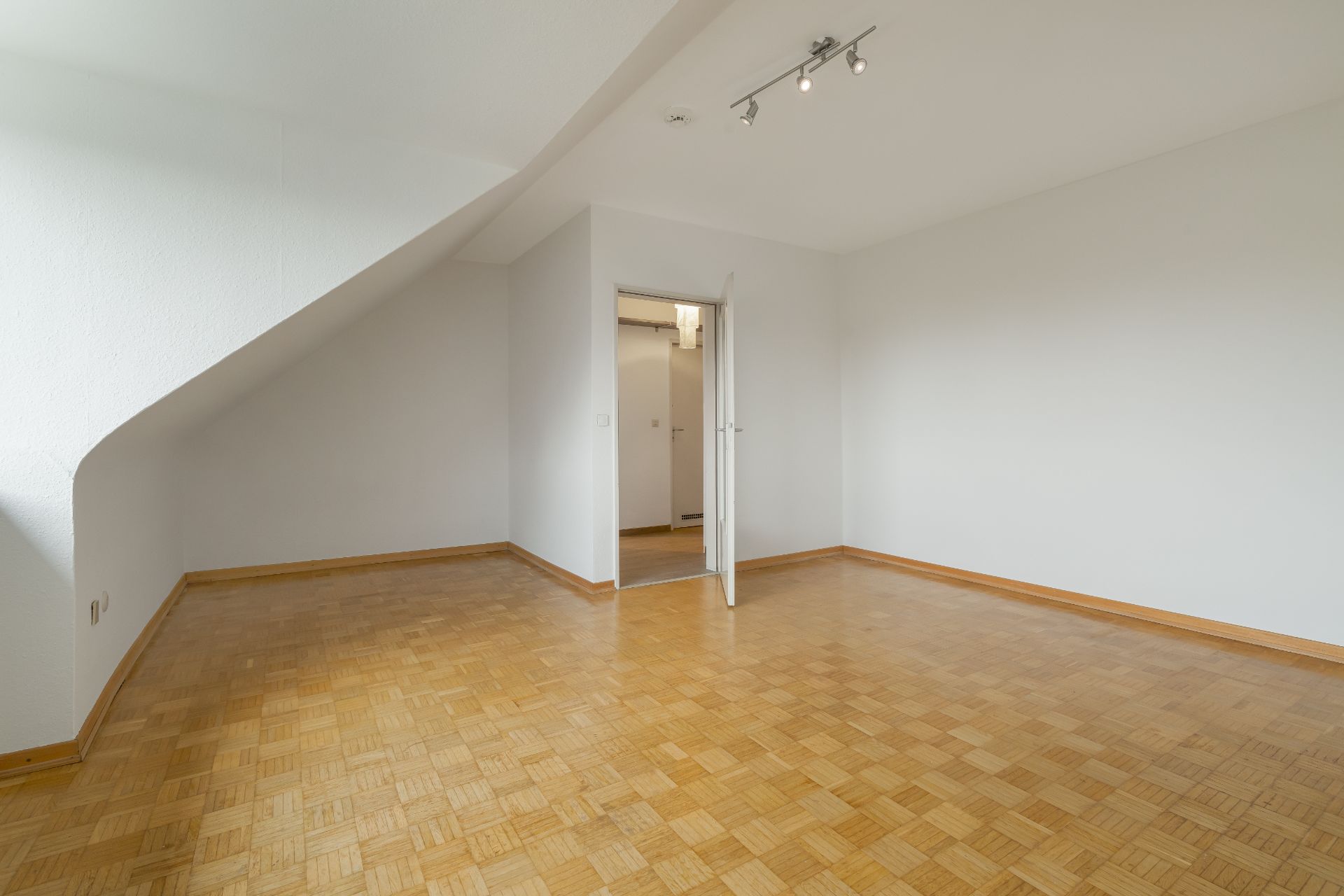 Leerer Raum mit Parkettboden, weißen Wänden und einer Tür in der Mitte.