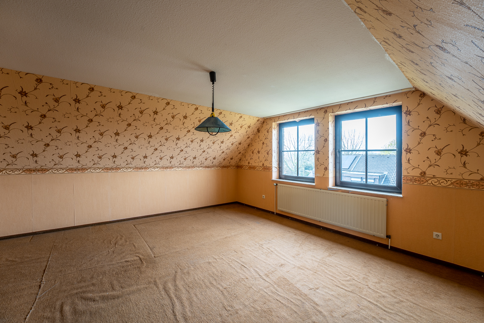 Leeres Zimmer mit gemusterter Tapete, zwei Fenstern und Lampe an der Decke.