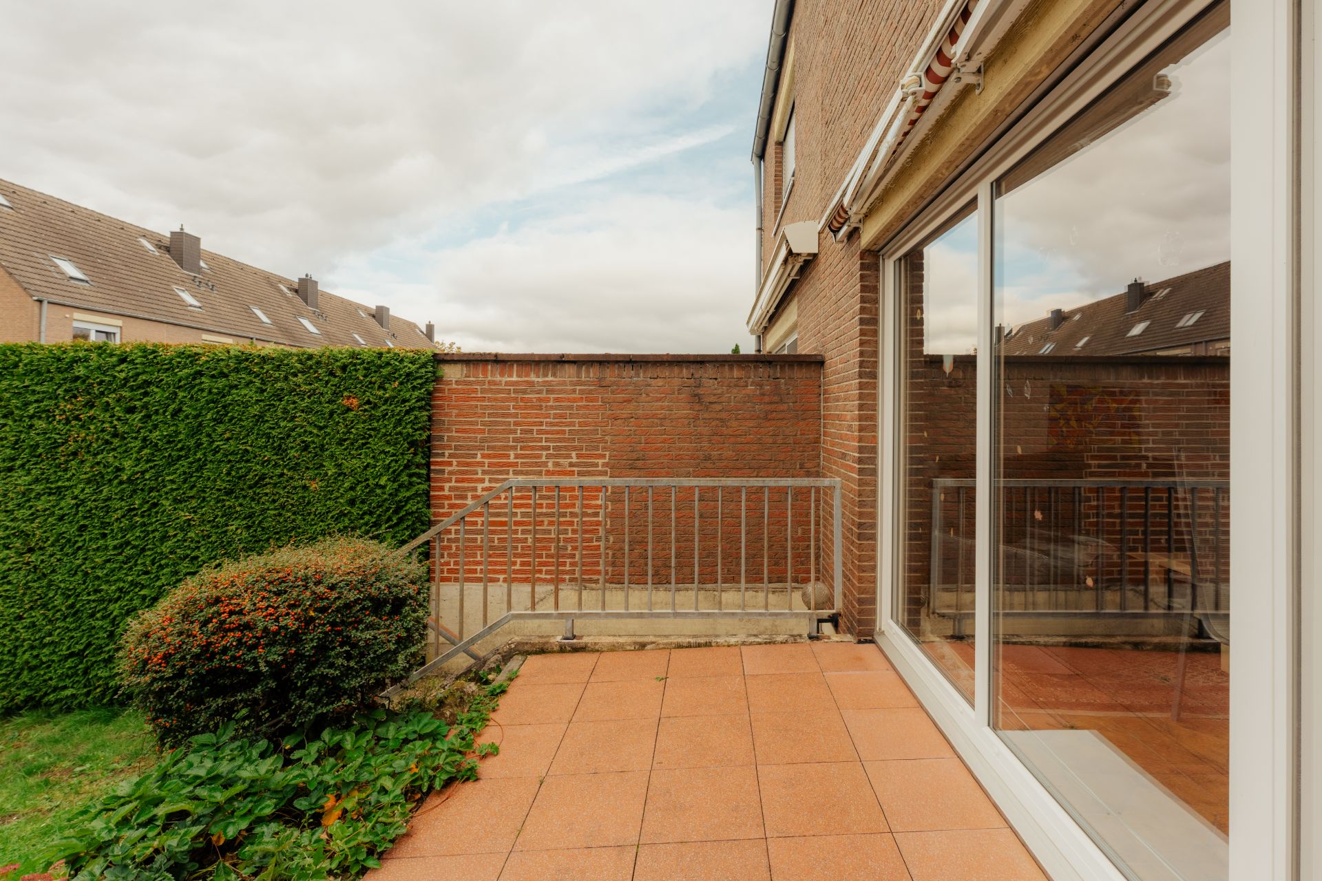 Terrasse mit Fliesenboden, Glas-Schiebetür, Hecke und Backsteinmauer.