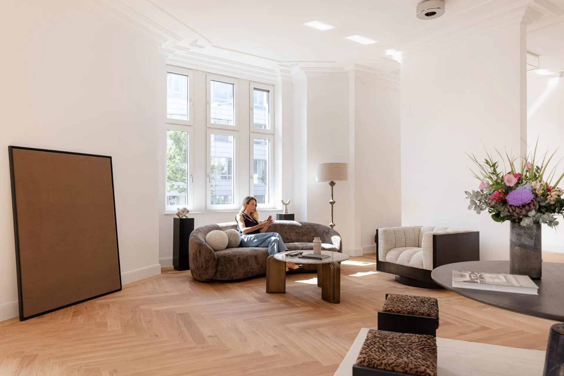 Helles Wohnzimmer mit großen Fenstern, modernen Möbeln und einer sitzenden Person auf dem Sofa.