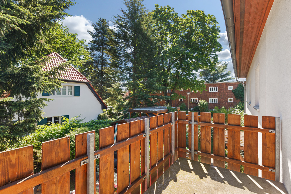 Balkon mit Holzgeländer, Blick auf Bäume und Nachbarhäuser bei sonnigem Wetter.