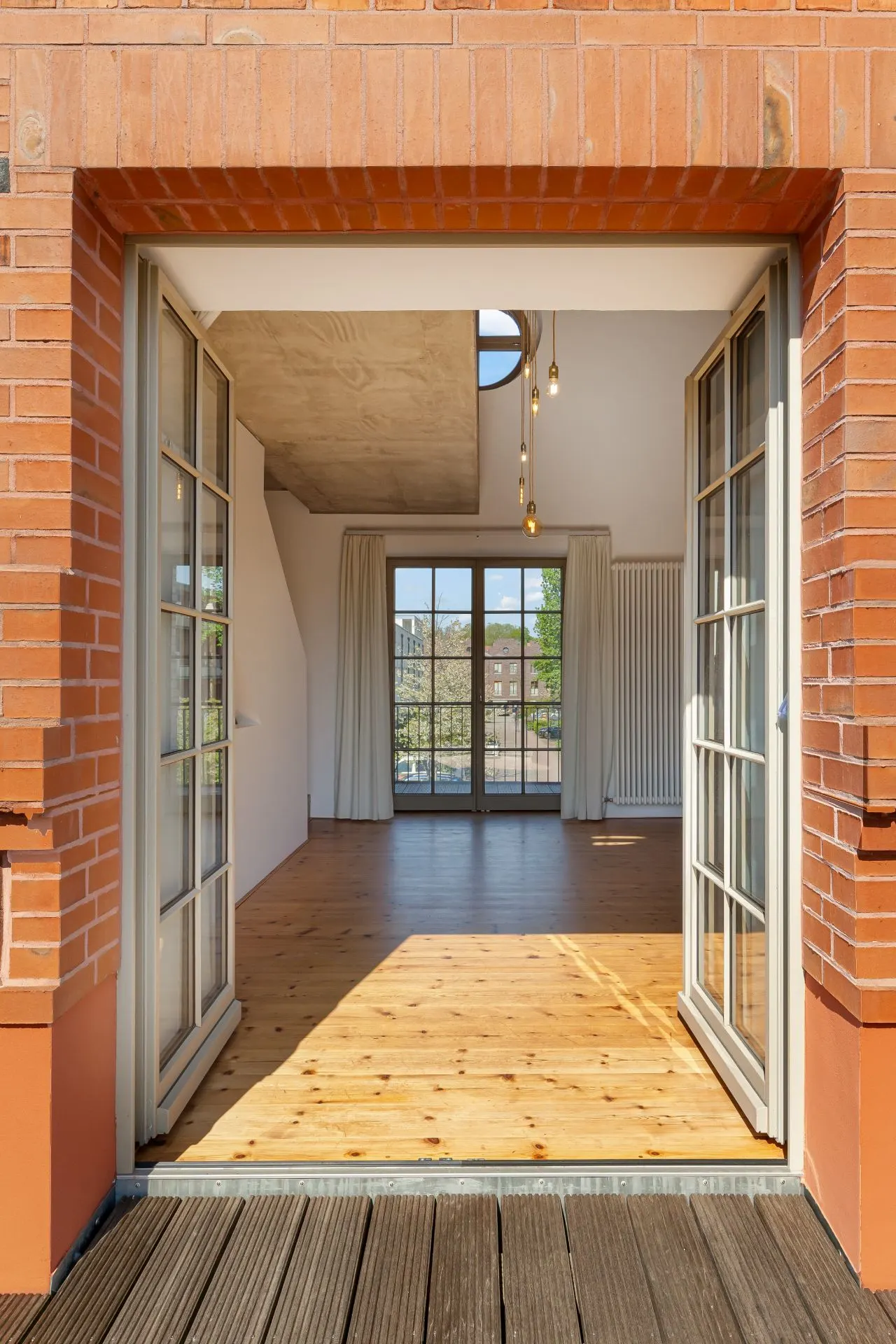 Blick durch geöffnete Glastüren in ein helles Zimmer mit Holzfußboden und großem Fenster.