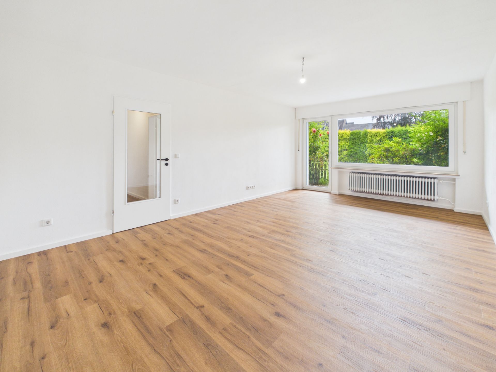 Helles, leeres Wohnzimmer mit Holzfußboden, weißer Tür und großem Fenster mit Blick ins Grüne.