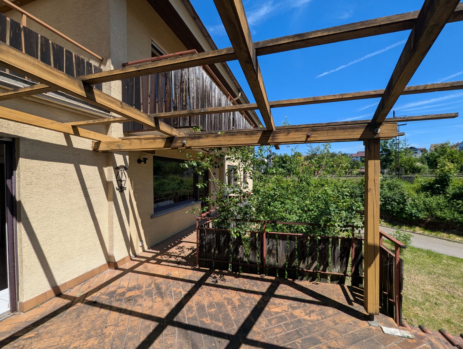 Sonnige Terrasse mit Holzpergola, Pflanzenkübel und Holzbalkon, angrenzend an einen Garten.