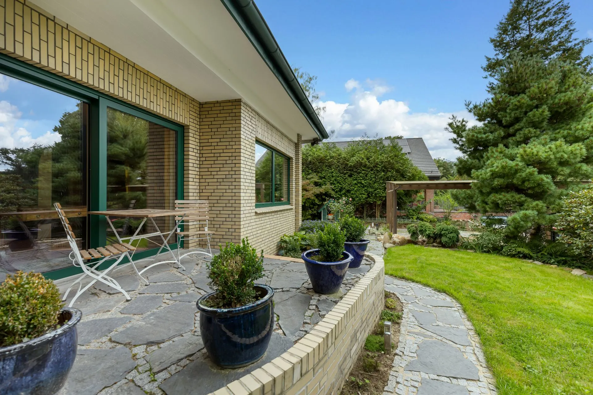Steinterrasse mit Stühlen, Blumentöpfen und Blick auf Garten neben einem Wohnhaus.