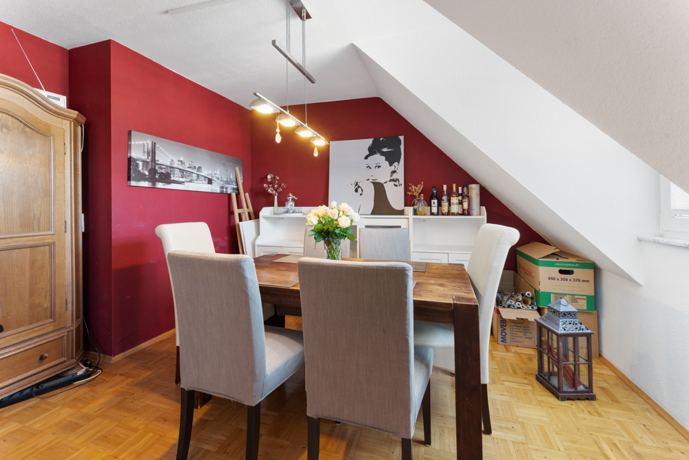 Esszimmer mit Holztisch, vier Stühlen, roter Wand, Blumen und Umzugskartons unter Schräge.