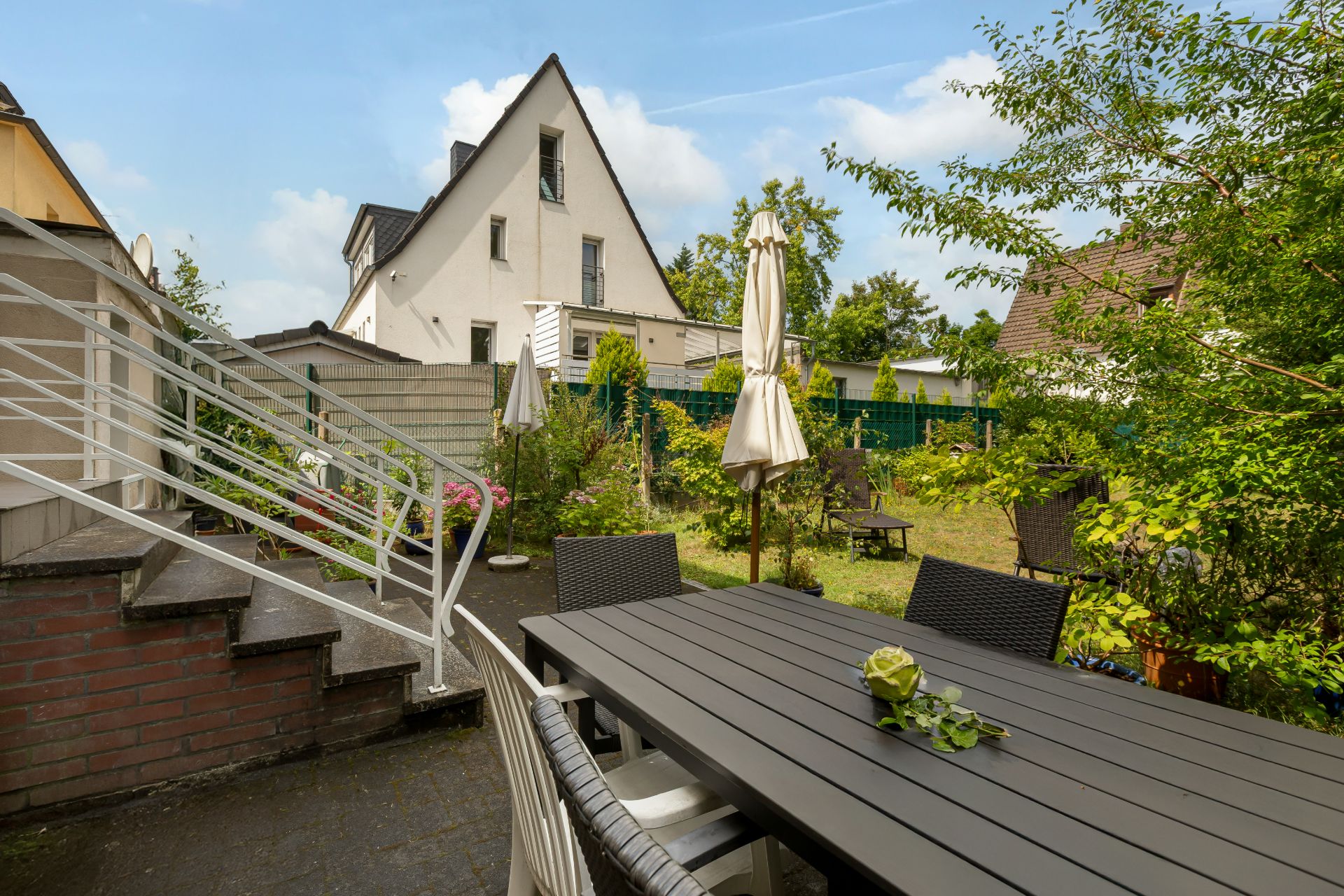 Garten mit Terrasse, Tisch und Stühlen, daneben Treppe, im Hintergrund Wohnhäuser und Bäume.