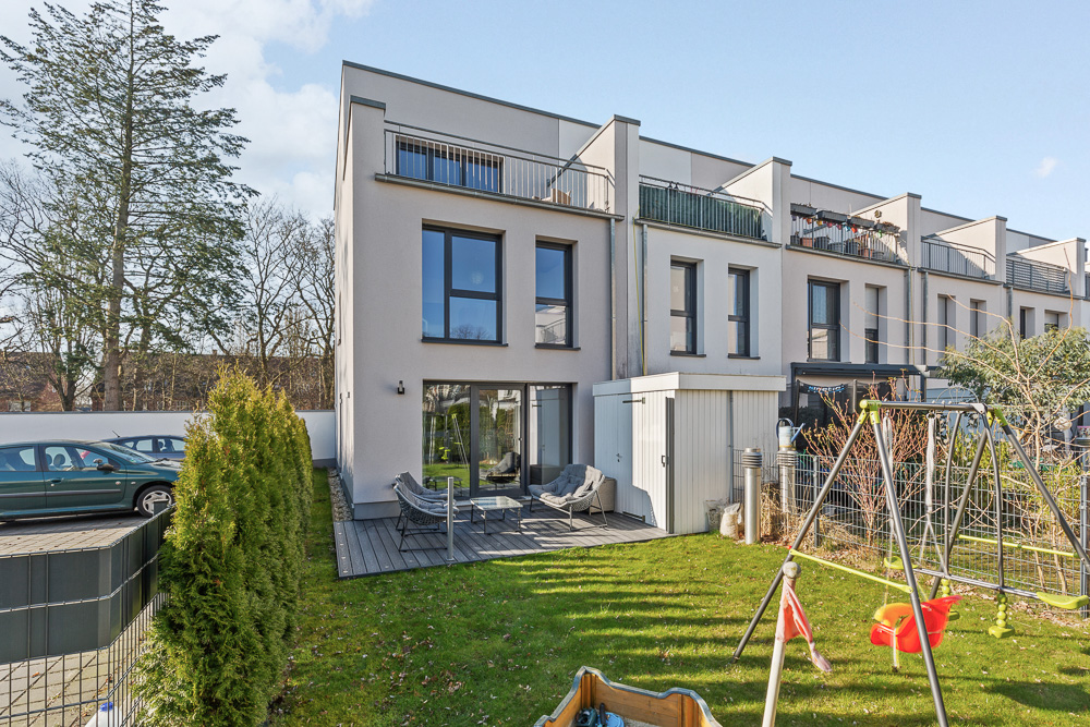 Modernes Reihenhaus mit Garten, Terrasse, Gartenhäuschen und Spielplatz mit Schaukel im Vordergrund.